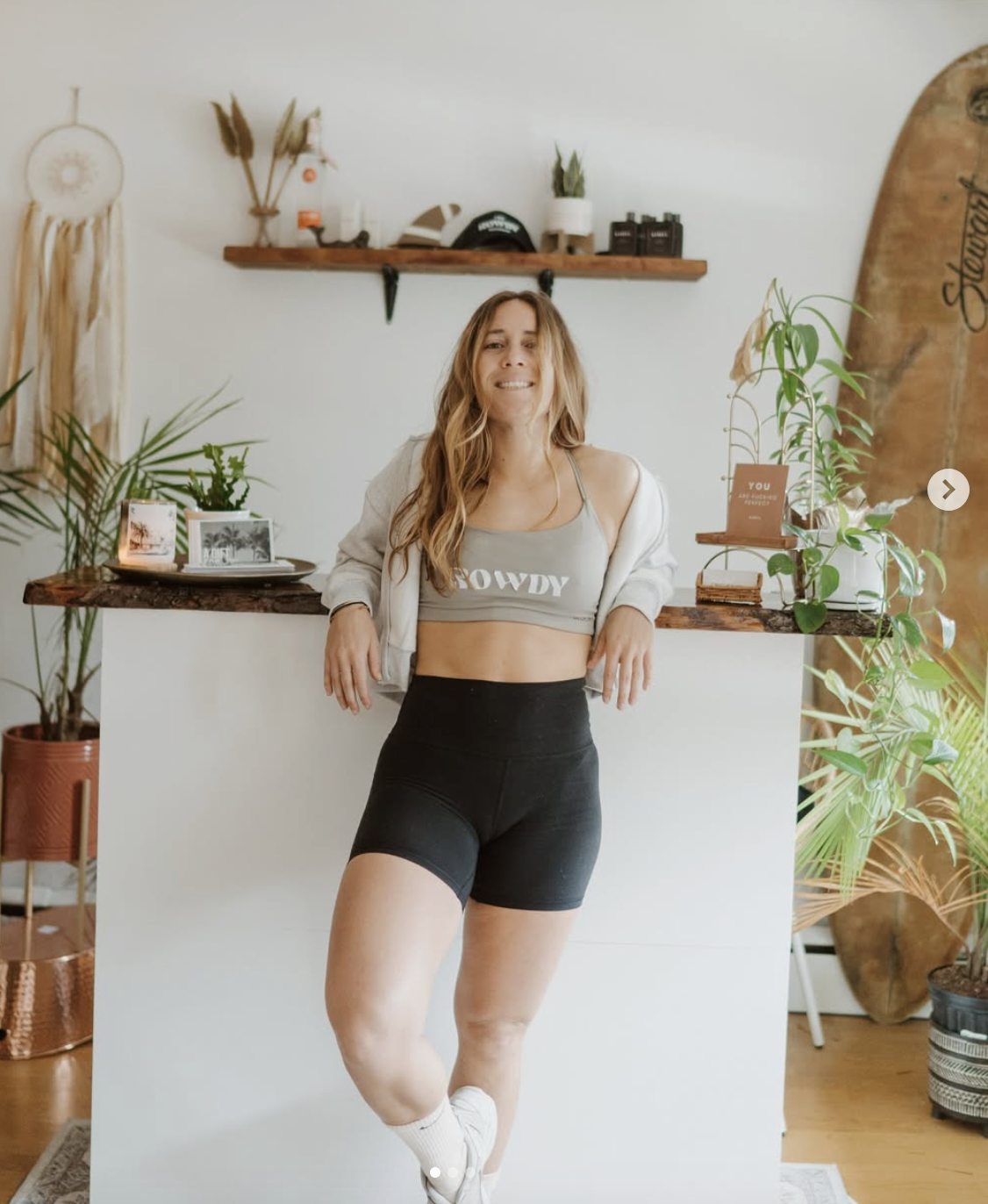 A woman with long wavy hair wearing black shorts, a gray crop top with the word 'COWDY', and a light-colored jacket hanging off her shoulders, standing in a room with plants and decor items on shelves and a counter.