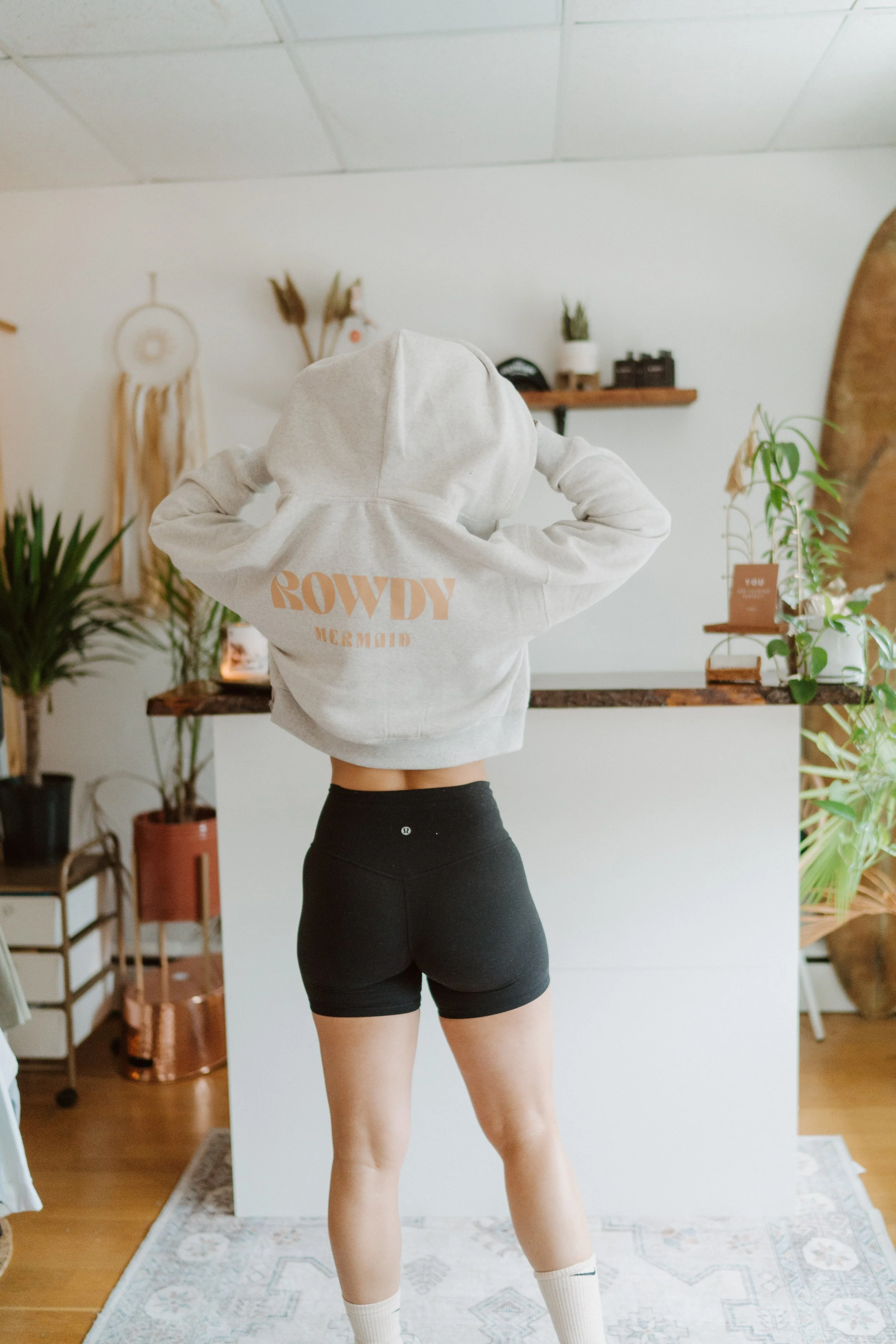 Person standing indoors wearing a white hoodie with text on the back, black shorts, and white socks, with plants and decorative items visible in the background.