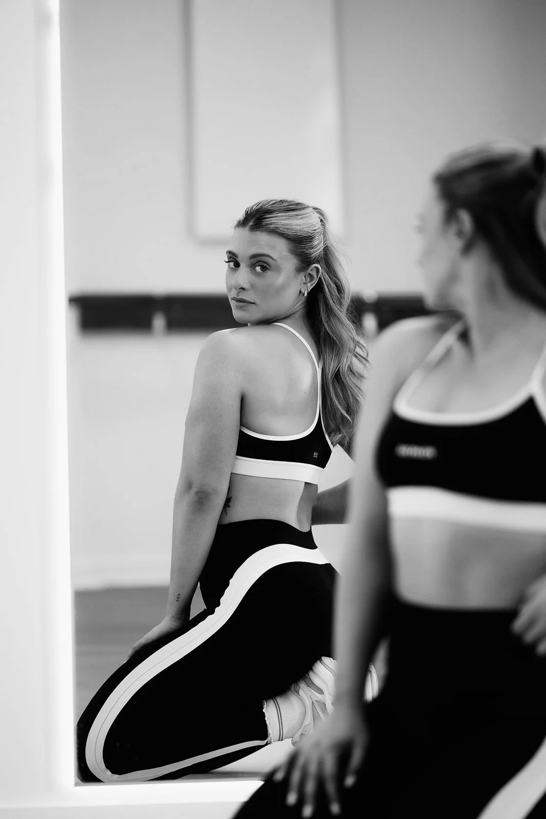 Two women in sports bras and leggings in a gym, one looking at the mirror and the other partially visible, both sitting on their heels.