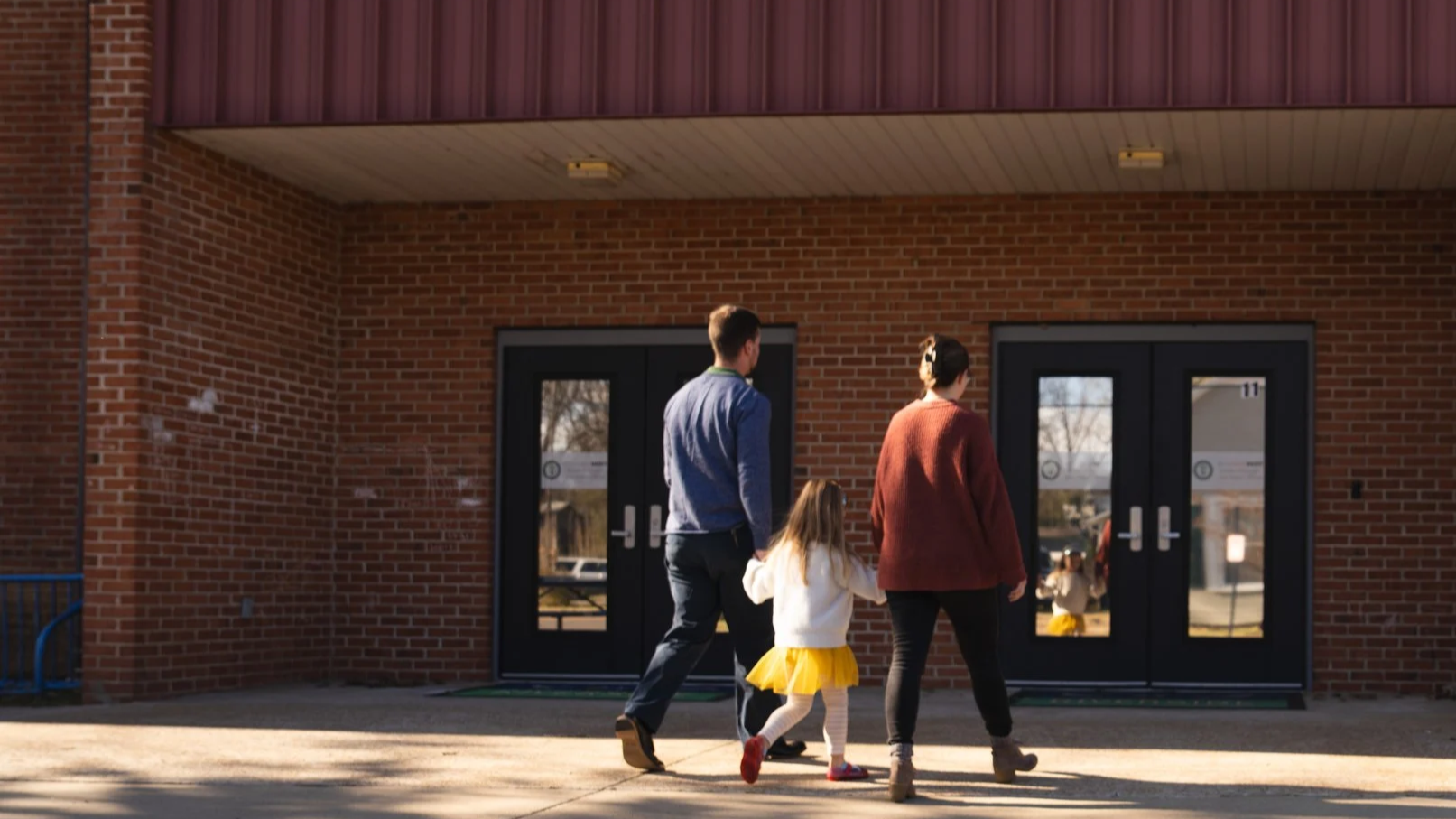 Family walking out of a school building with brick walls and glass double doors on a sunny day.