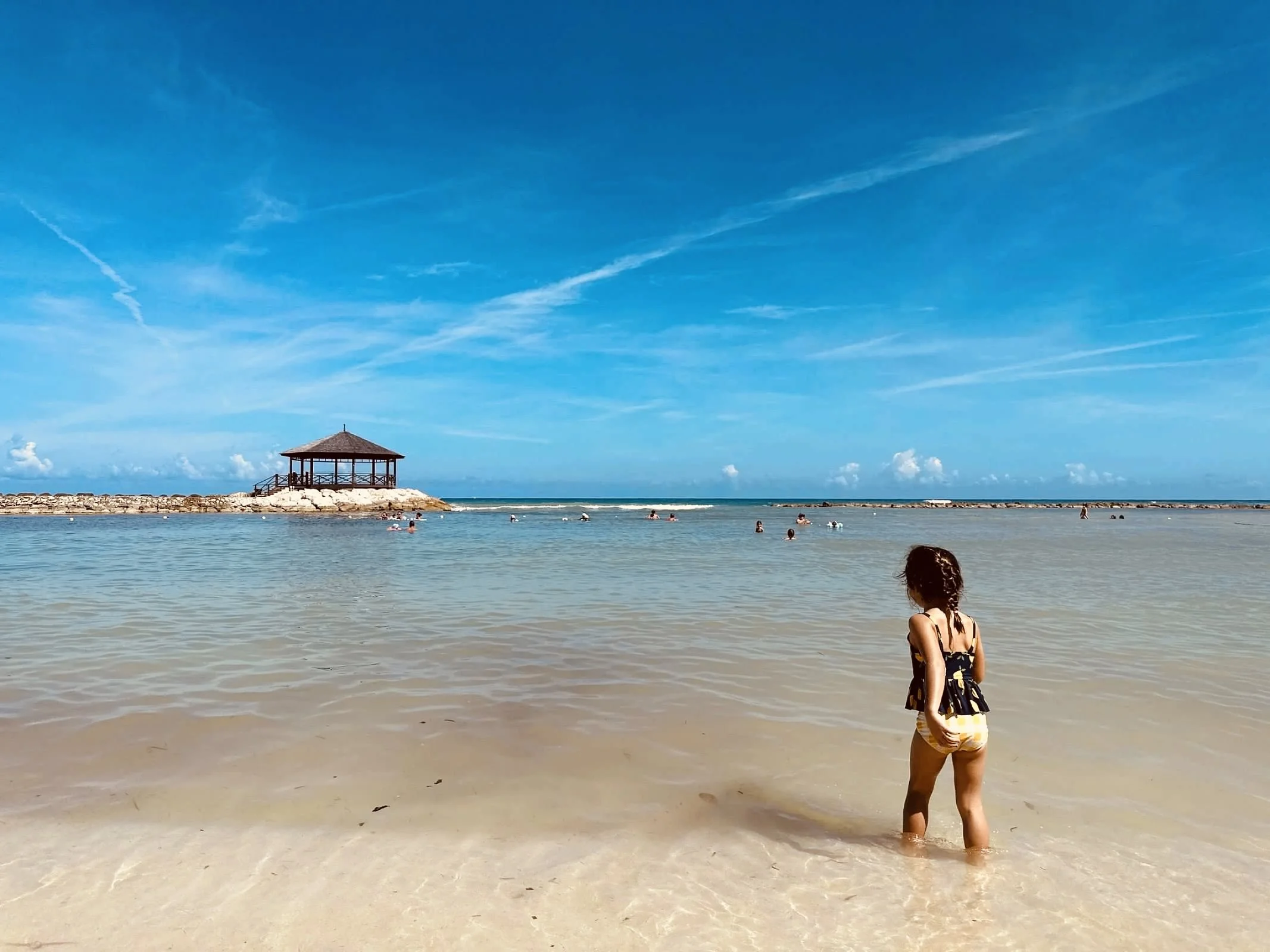 A child stands in shallow water on a beach, facing a wooden gazebo on a rocky jetty under a blue sky.