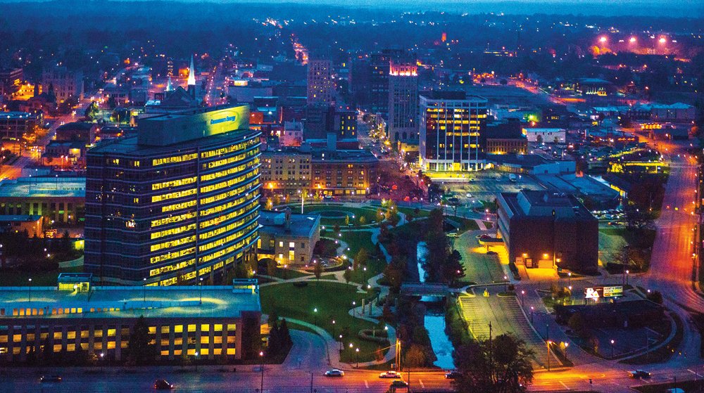 Nighttime cityscape with illuminated buildings, streets, and a river running through a park.