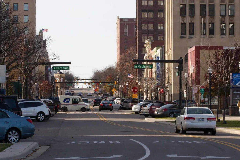 City street intersection with parked cars, buildings, traffic lights, and street signs for N Mechanic and S Mechanic.