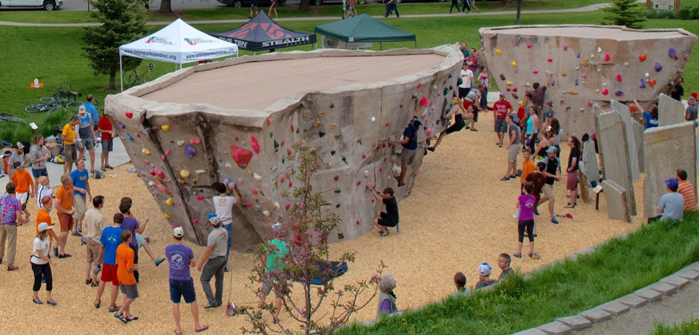 An outdoor rock climbing event with several people climbing and observing near large artificial climbing walls, set in a park with tents and grassy areas.