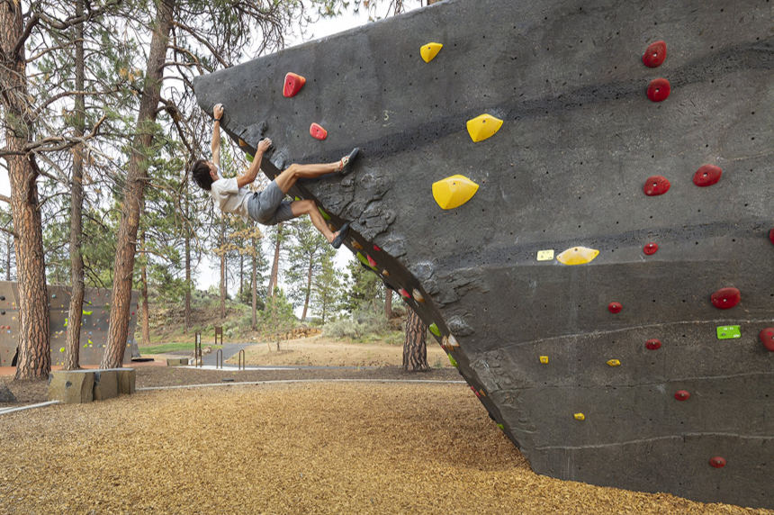 A person climbing an outdoor bouldering wall in a park with trees in the background.