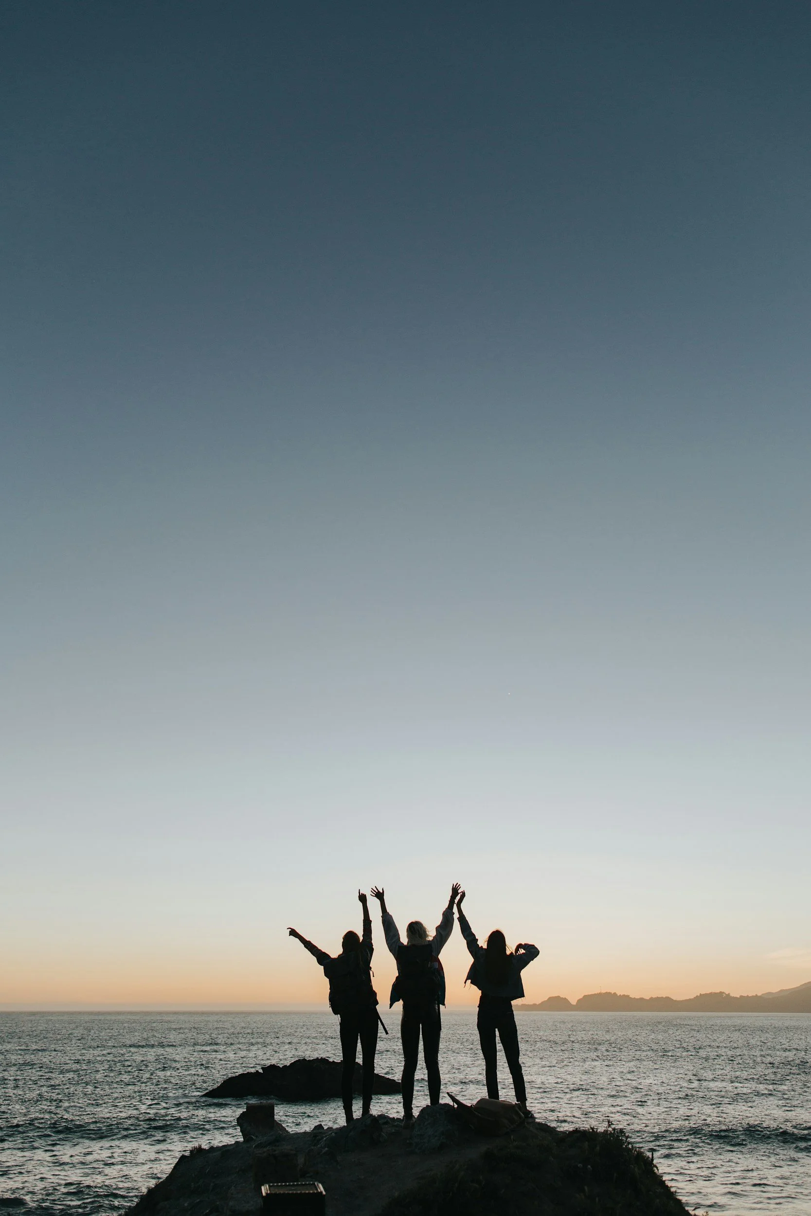 Four people standing on a rocky coastline at sunset with their arms raised, facing the ocean and silhouette of islands in the distance.