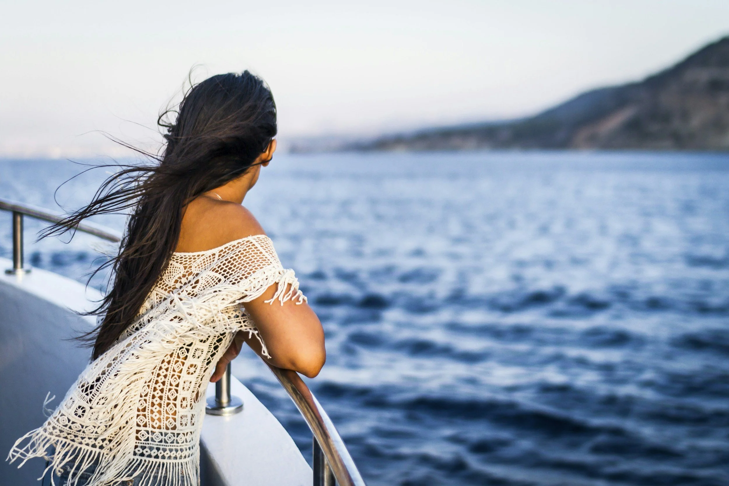A woman with long dark hair looking out over the ocean from a boat, wearing a white crochet top, with a hilly coastline in the distance.