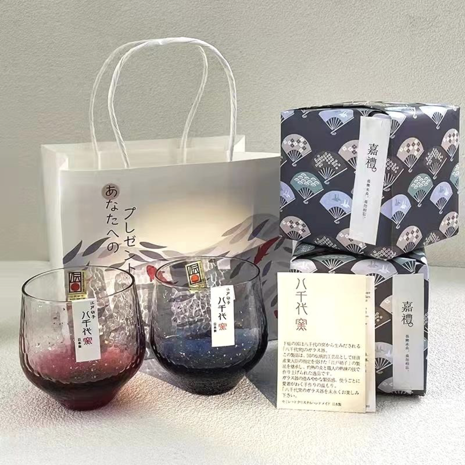 Two glasses with red and blue liquids, decorative labels, origami paper boxes with traditional Japanese pattern, a white gift bag with Japanese writing, and a paper note with Japanese text.