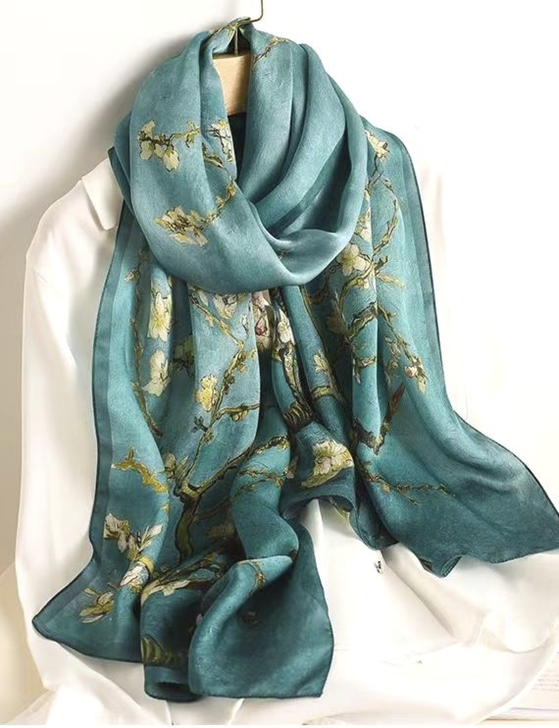 A teal silk scarf with a floral pattern, draped over a white garment, hanging on a wooden hanger.