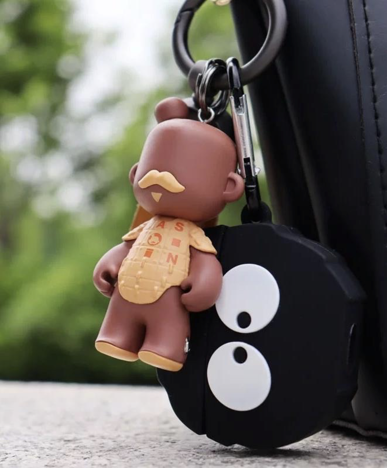 A keychain with a plush toy of a bald man with a mustache and beige outfit, hanging from a backpack strap, with a black case featuring a white cartoon face design attached.
