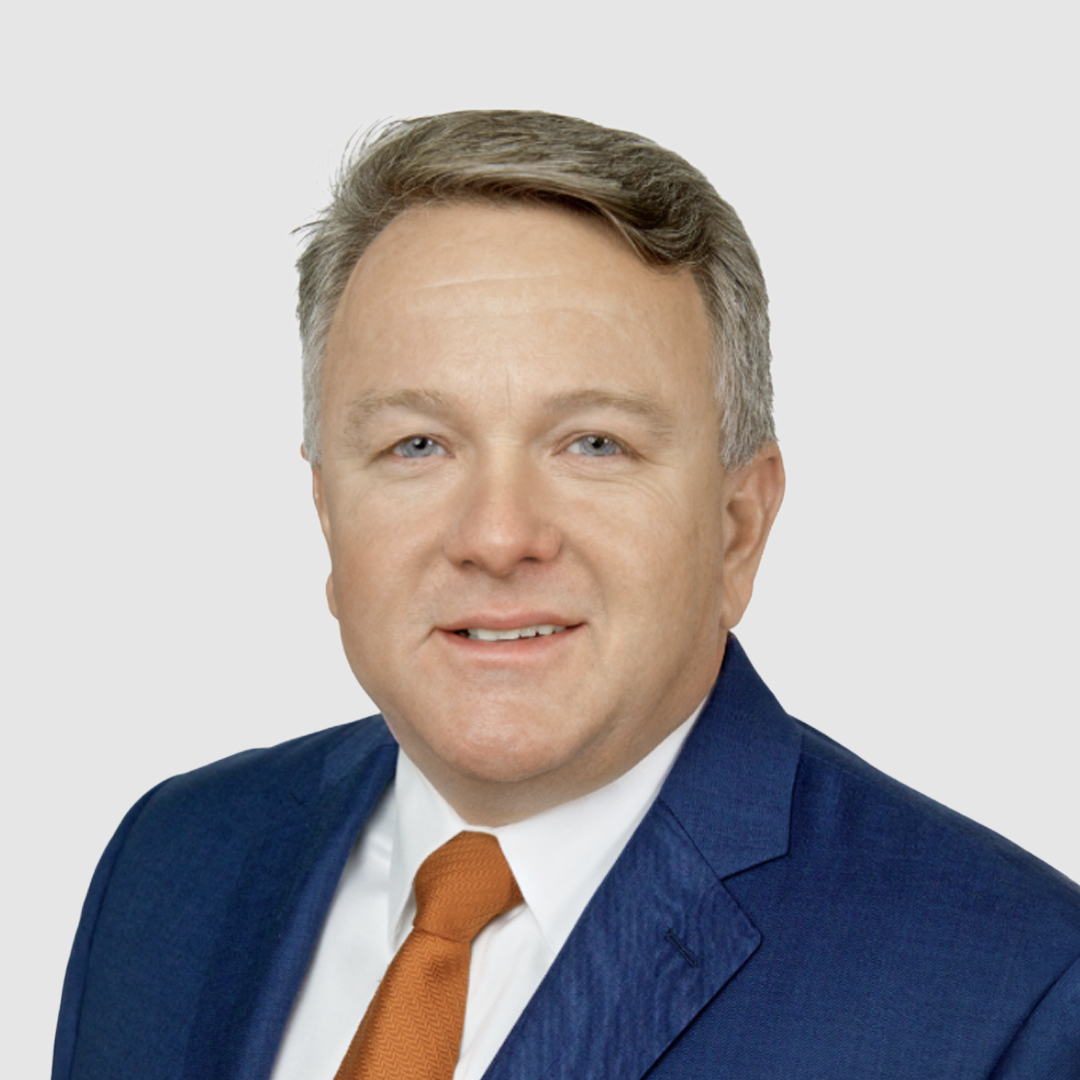 A professional headshot of a middle-aged man with short, light brown hair, wearing a navy blue suit, white shirt, and orange tie, against a plain white background.