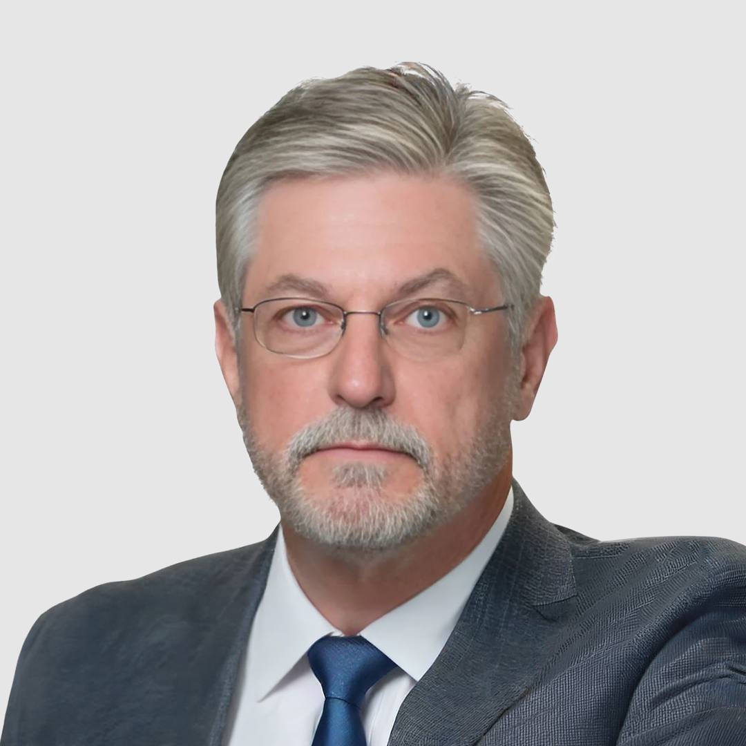 Close-up of a middle-aged man with gray hair, beard, and glasses, wearing a suit against a white background.