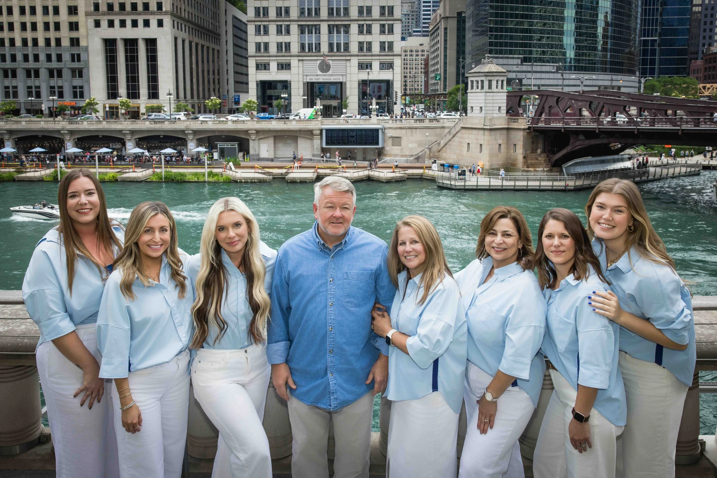 A group of eight people, seven women and one man, standing together outdoors on a boardwalk by a body of water in an urban area with tall buildings and a bridge in the background.