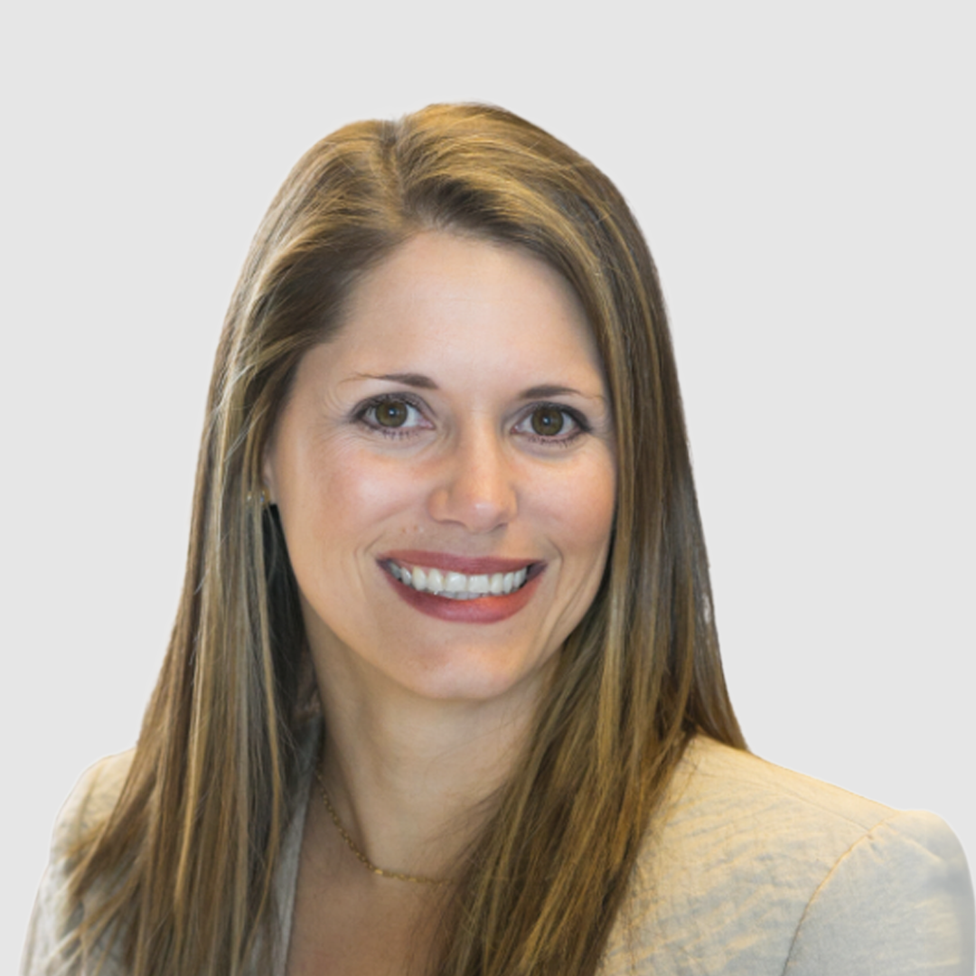 A woman with long light brown hair, smiling, wearing a beige blazer and a thin gold necklace against a white background.