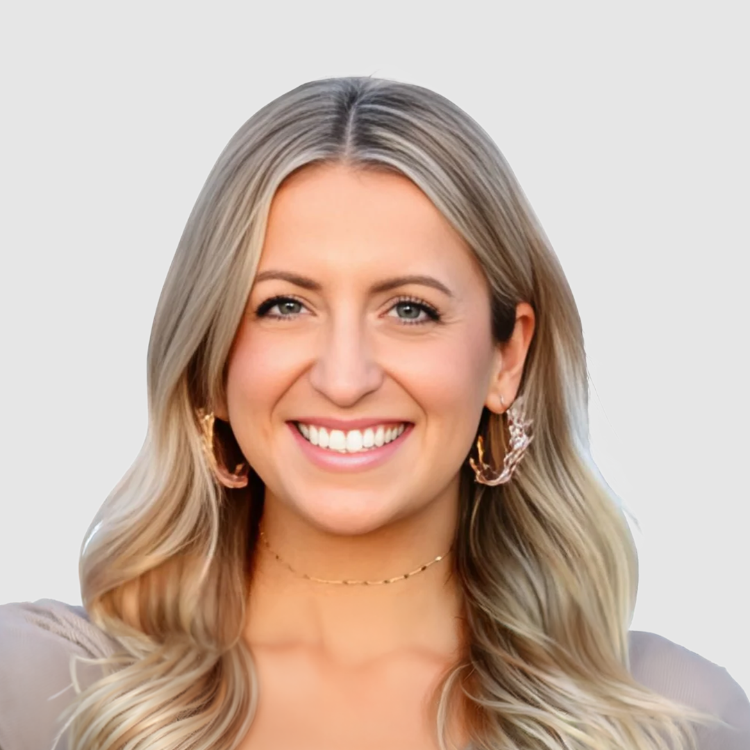 Close-up of a woman with blonde hair, blue eyes, and a bright smile, wearing gold earrings and a delicate necklace.