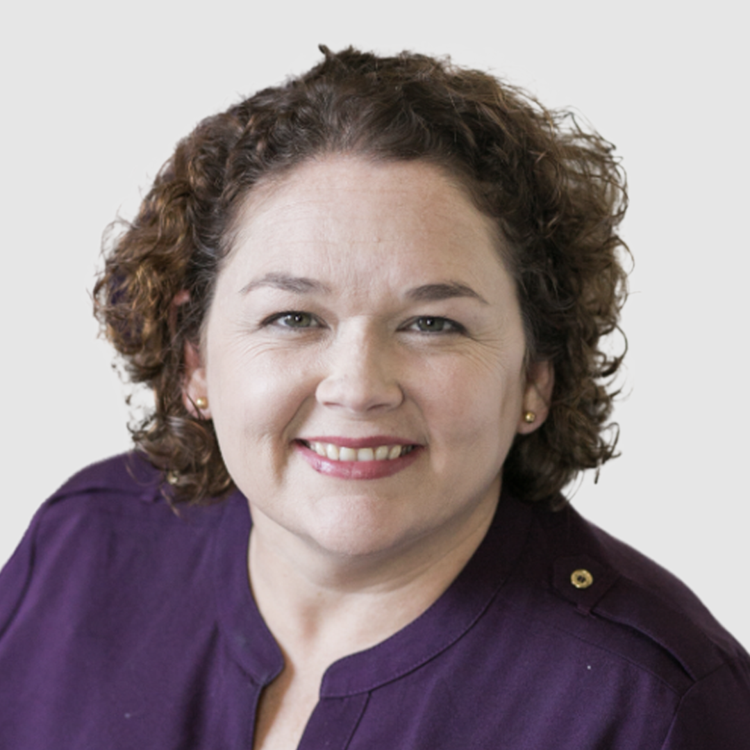 Portrait of a woman with shoulder-length curly brown hair, wearing a purple top, smiling against a plain white background.
