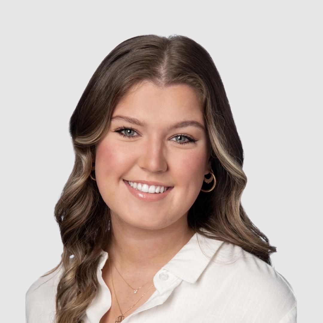 Young woman with long wavy brown hair, wearing a white button-up shirt, gold jewelry, and earrings, smiling against a plain white background.