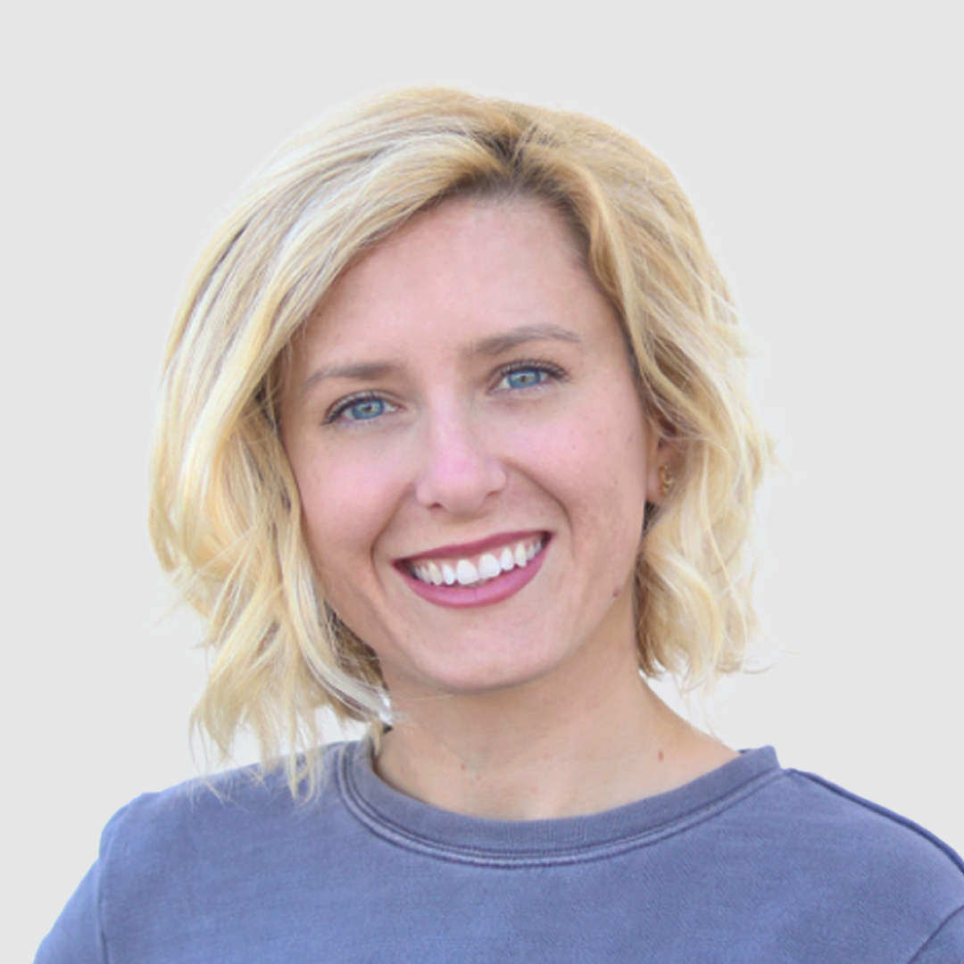 A smiling woman with blonde hair and blue eyes, wearing a gray shirt, against a light background.