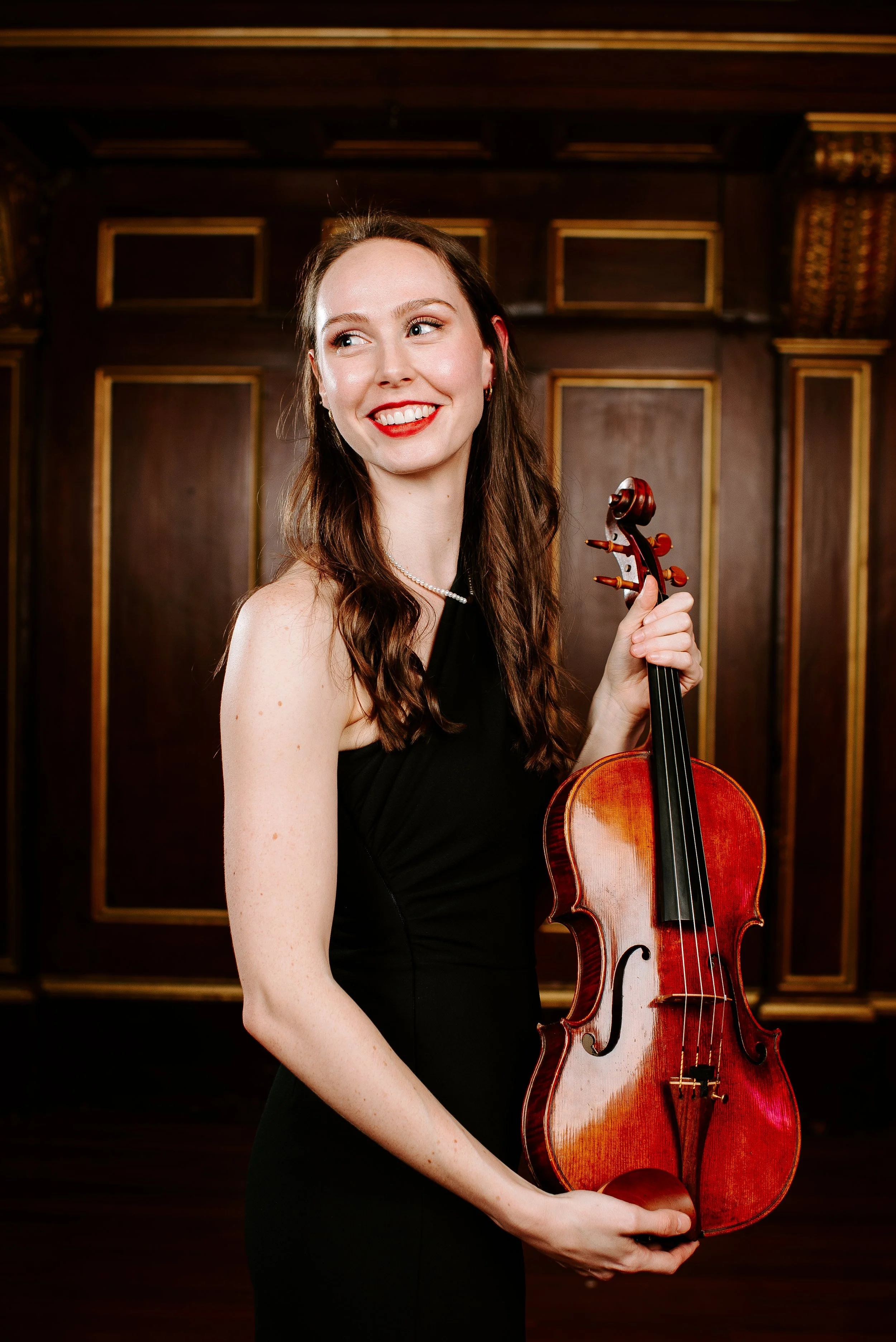 A young woman with long brown hair, wearing a black sleeveless dress and a pearl necklace, holding a wooden violin in front of a dark wood-paneled background with gold accents, smiling and looking to the side.