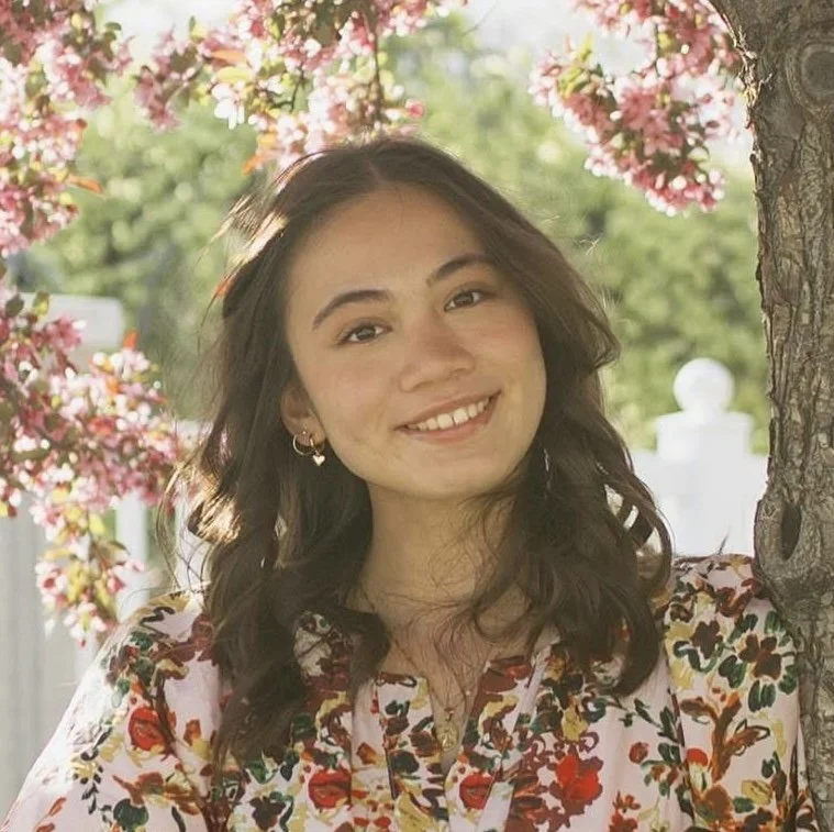 A young woman with wavy dark hair and earrings smiling outdoors near a blossoming tree with pink flowers, green foliage, and a white fence in the background.