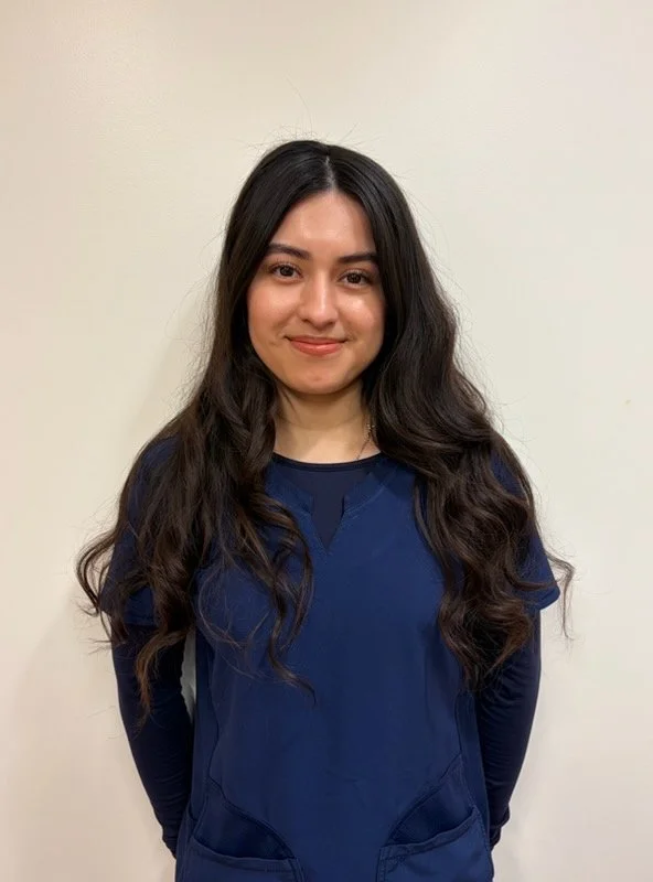 A woman with long dark hair wearing purple scrubs standing against a plain off-white wall.