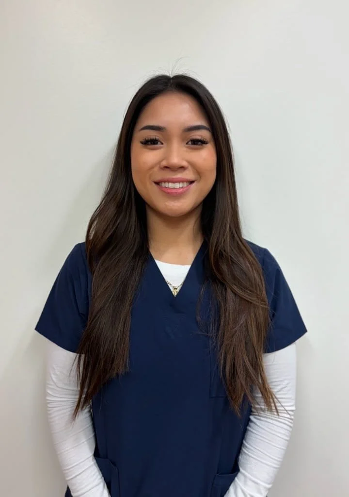 A young medical assistant with dark hair tied back, smiling, wearing gold hoop earrings.