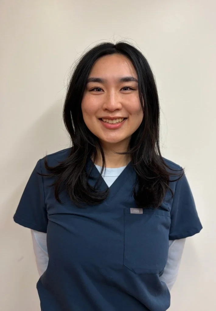 Young medical assistant with long black hair smiling at the camera in an office setting.