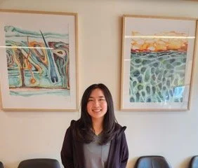 Young medical assistant with long black hair smiling at the camera in an office setting.