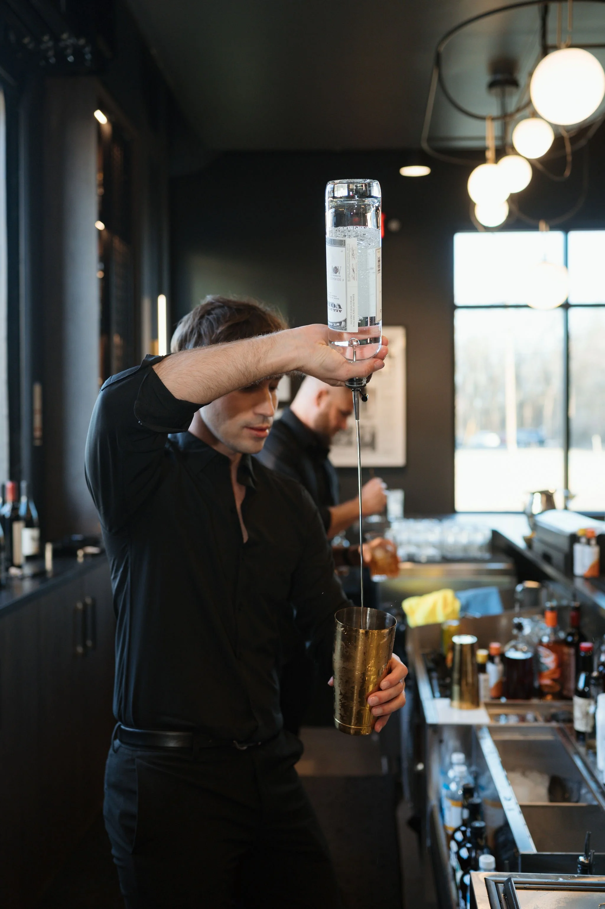 A bartender is pouring a clear liquid from a tall bottle into a golden shaker in a bar setting. The bar has dark walls, modern lighting, and large windows with natural light.