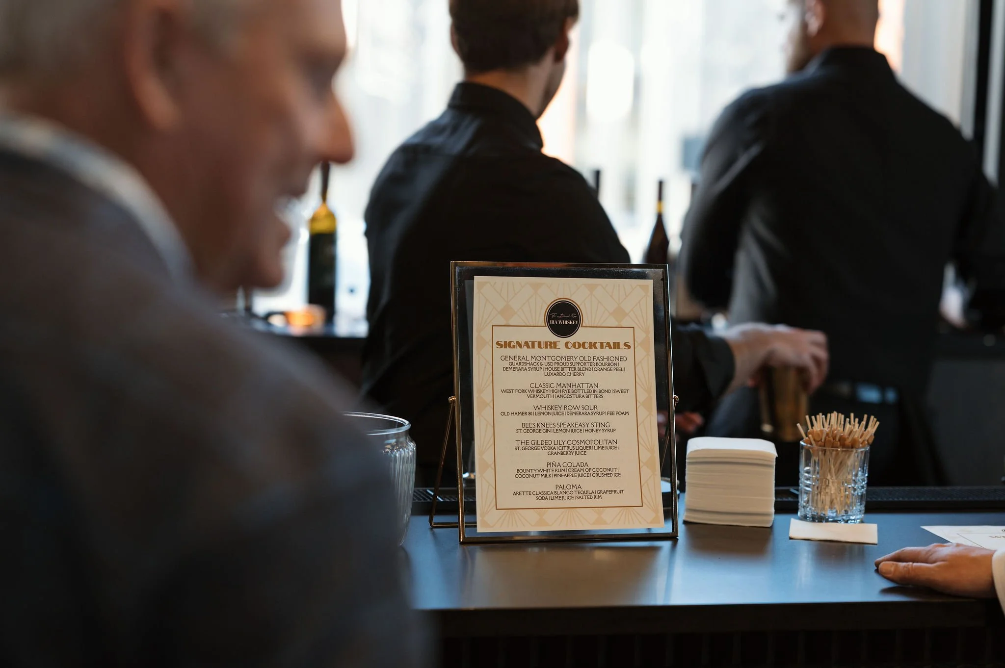 A bar or restaurant scene with a sign displaying a list of signature cocktails and a group of people sitting at the bar, with focus on a woman in the foreground and two men in the background.