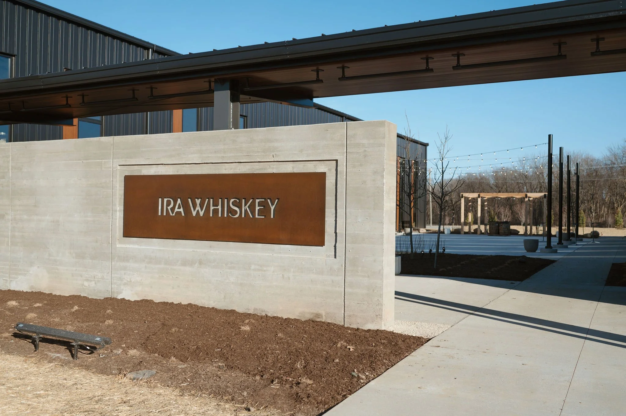 Modern building with a sign that reads 'IRAW Whiskey', a concrete wall, and outdoor seating with string lights and trees in the background.