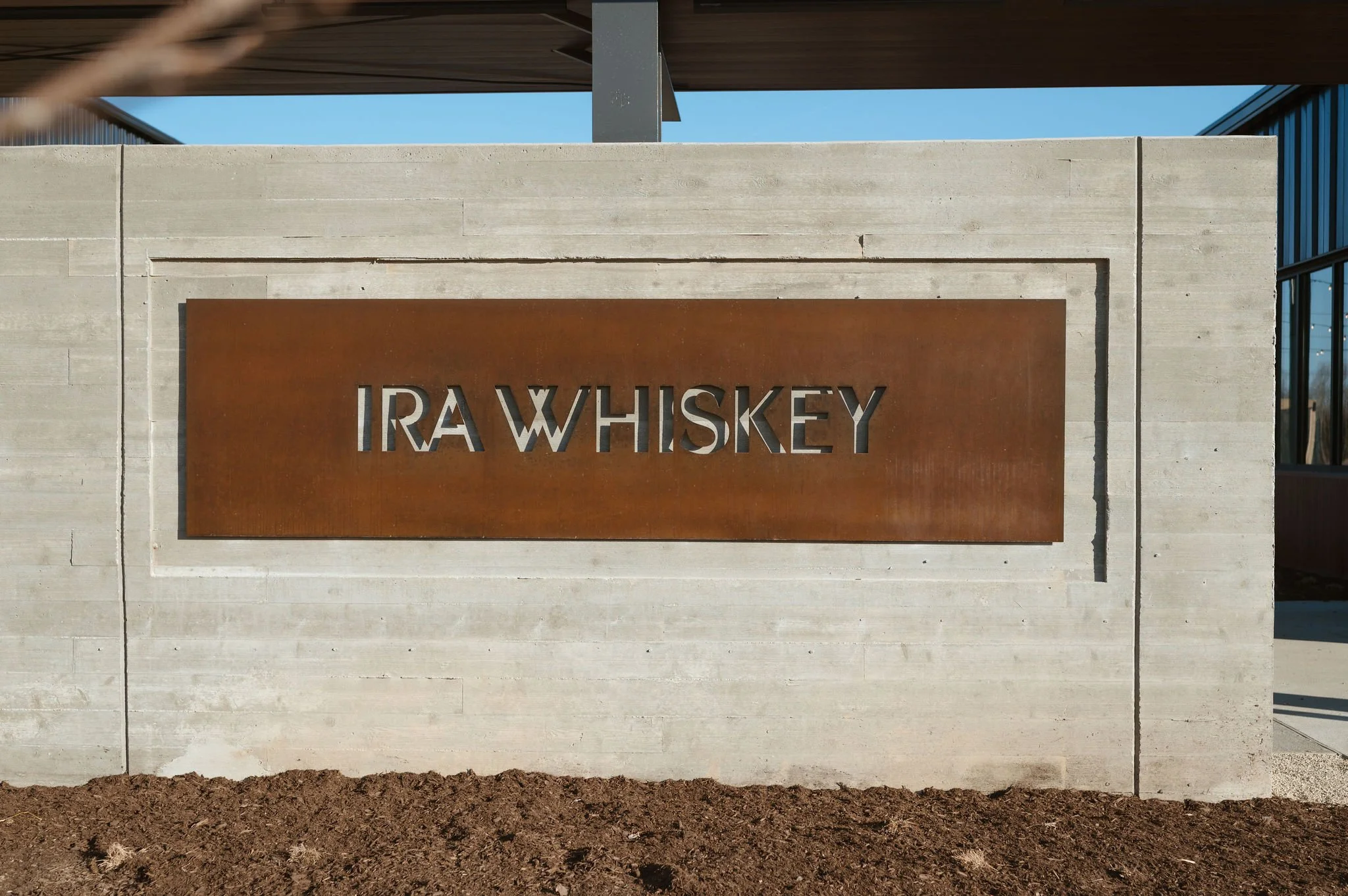 Sign that reads 'RA WHISKEY' mounted on a concrete wall with a modern building and blue sky in the background.