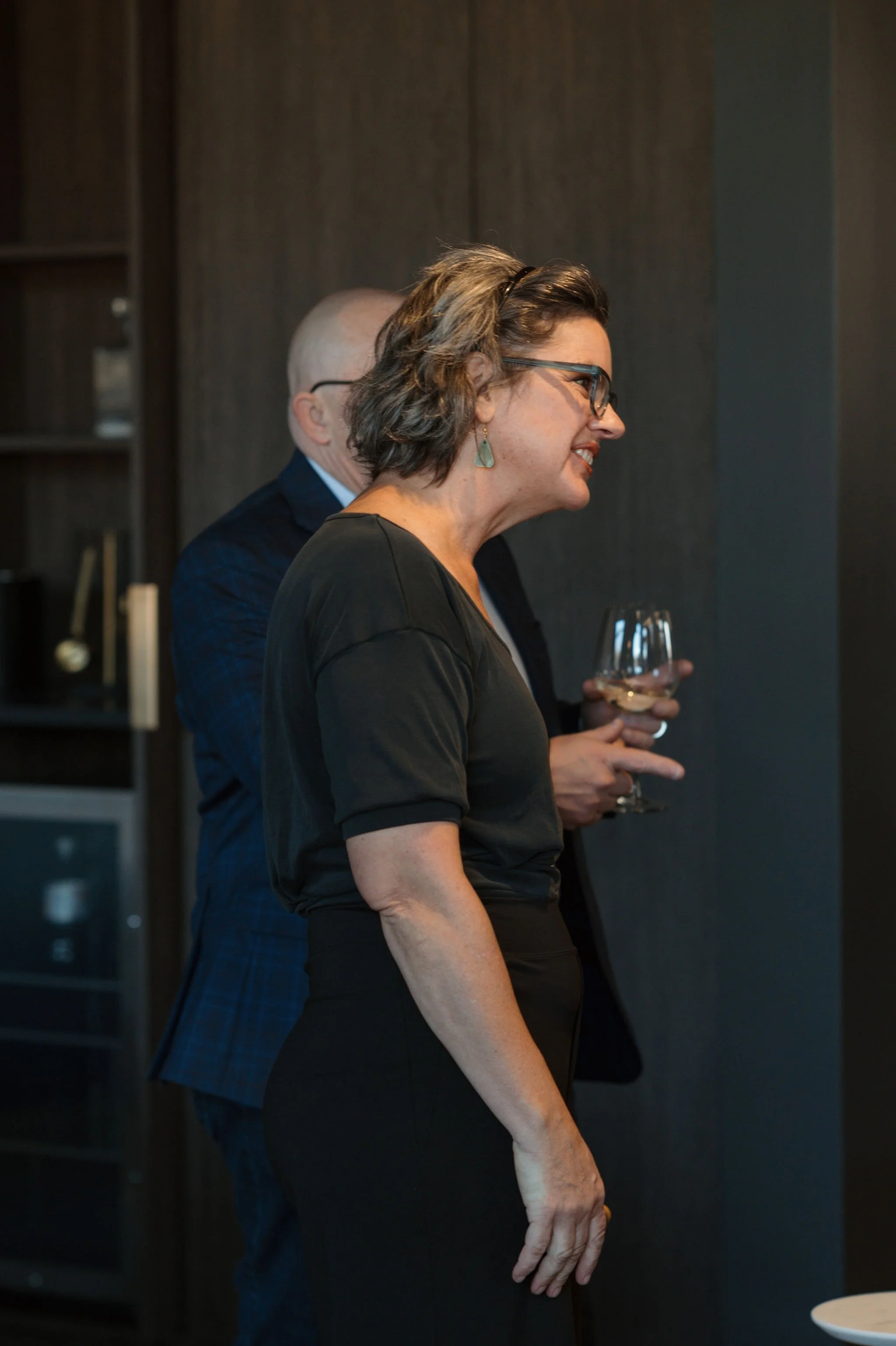 A woman wearing glasses with short wavy hair and earrings, holding a glass of white wine, standing next to a man in a dark suit and glasses. They are indoors with a dark wood background.