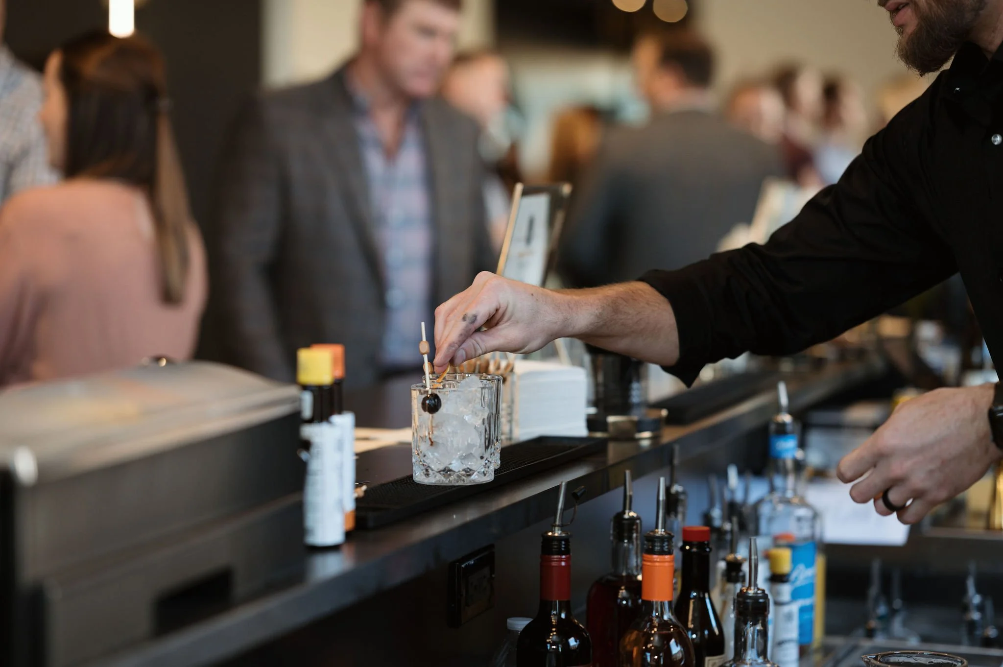 Bartender preparing a drink with alcohol bottles and mixing tools on a bar counter in a crowded setting.