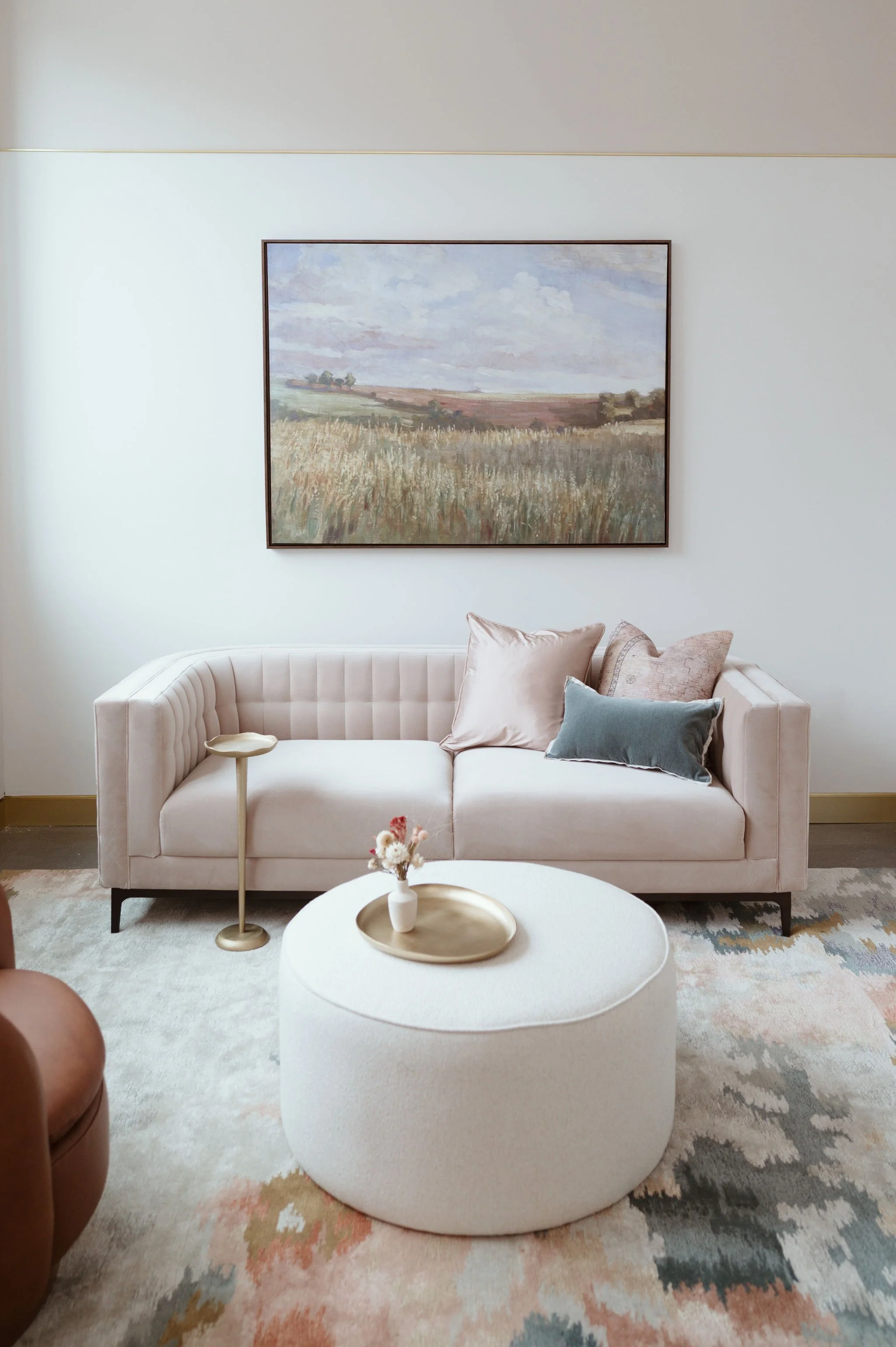 Living room with a light pink sofa with pillows, a cream-colored ottoman with a vase of flowers on it, a side table, and a large landscape painting on the wall. There is a patterned rug on the floor.