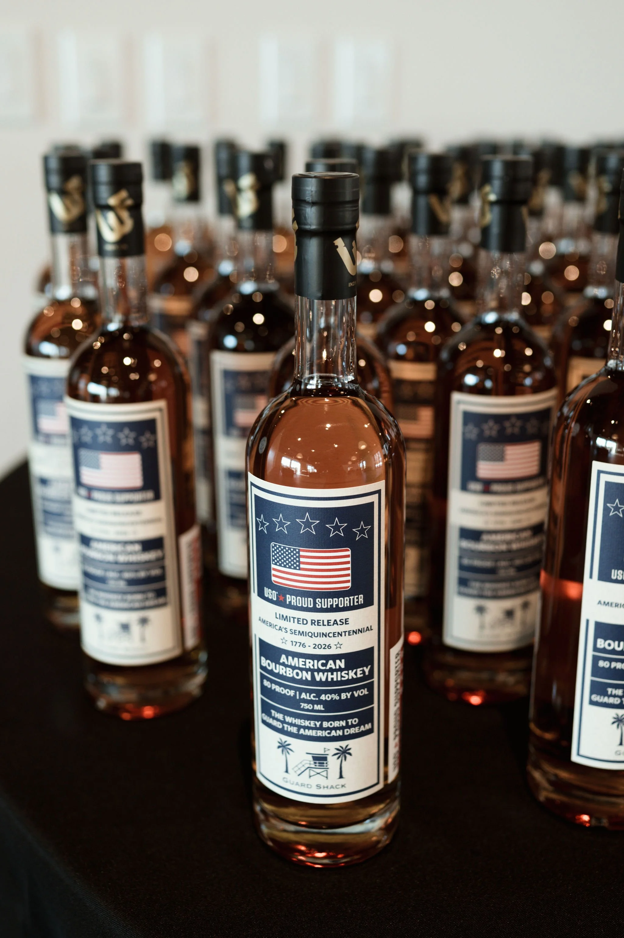 Rows of bottles of American Bourbon Whiskey with patriotic labels featuring the American flag and text celebrating America's semiquicentennial from 1776 to 2026, on a black surface.