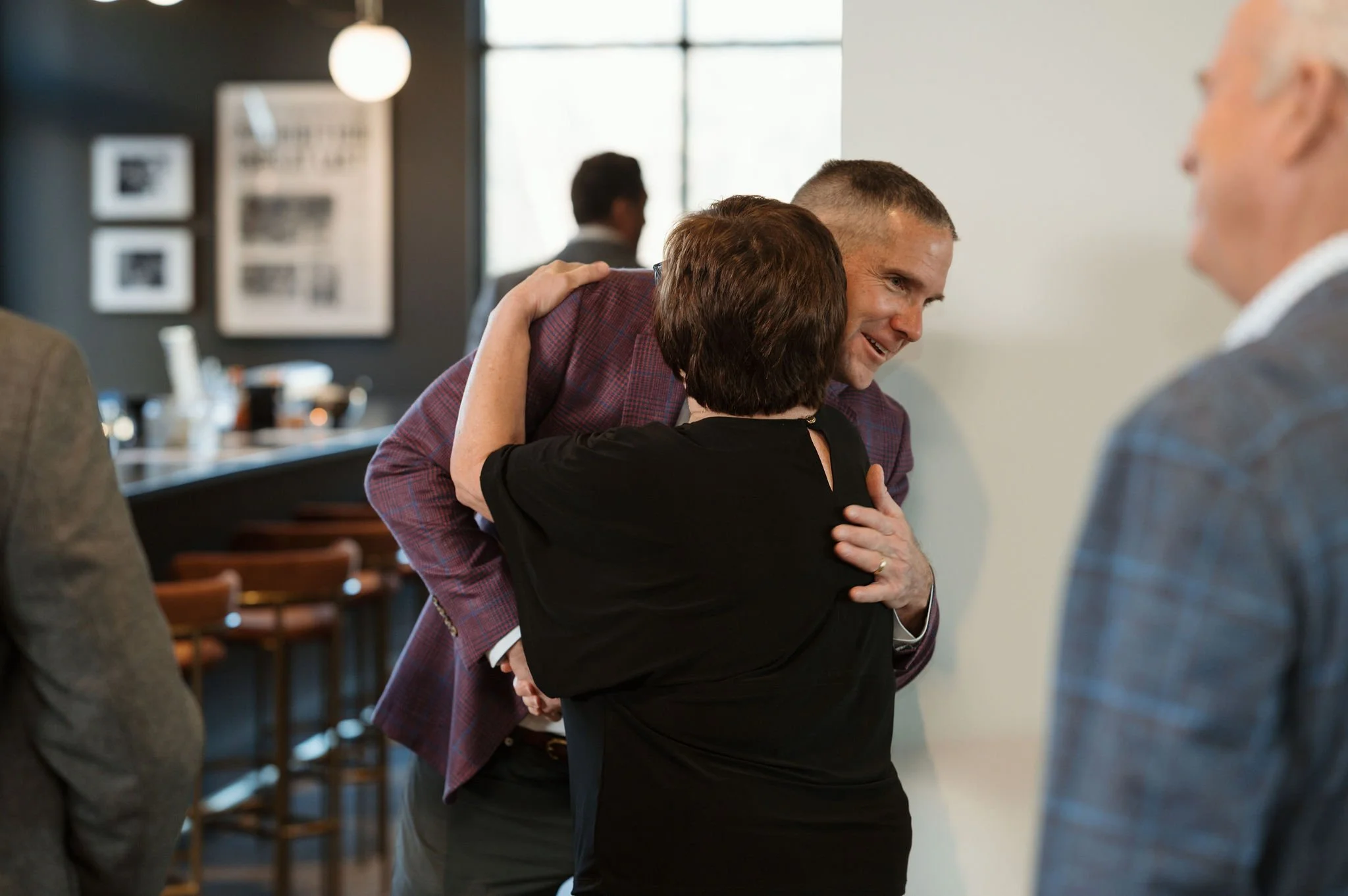 People are hugging in a friendly manner in a social gathering, likely in a restaurant or event space.