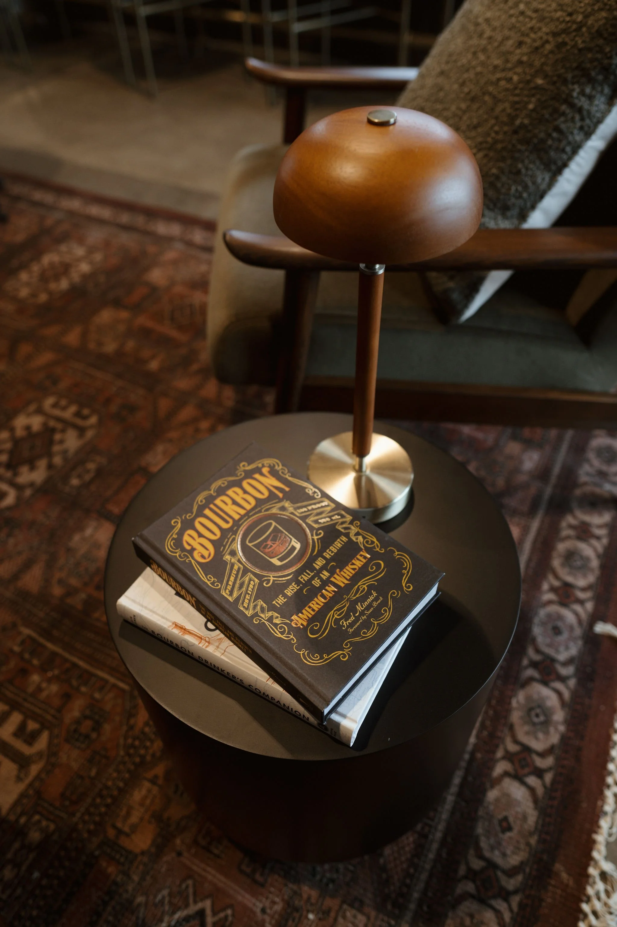 A round black side table with two books on it, one titled 'Gourbong' and the other 'Distillation Drinker'. A wooden armrest of a chair and a patterned carpet are visible in the background.