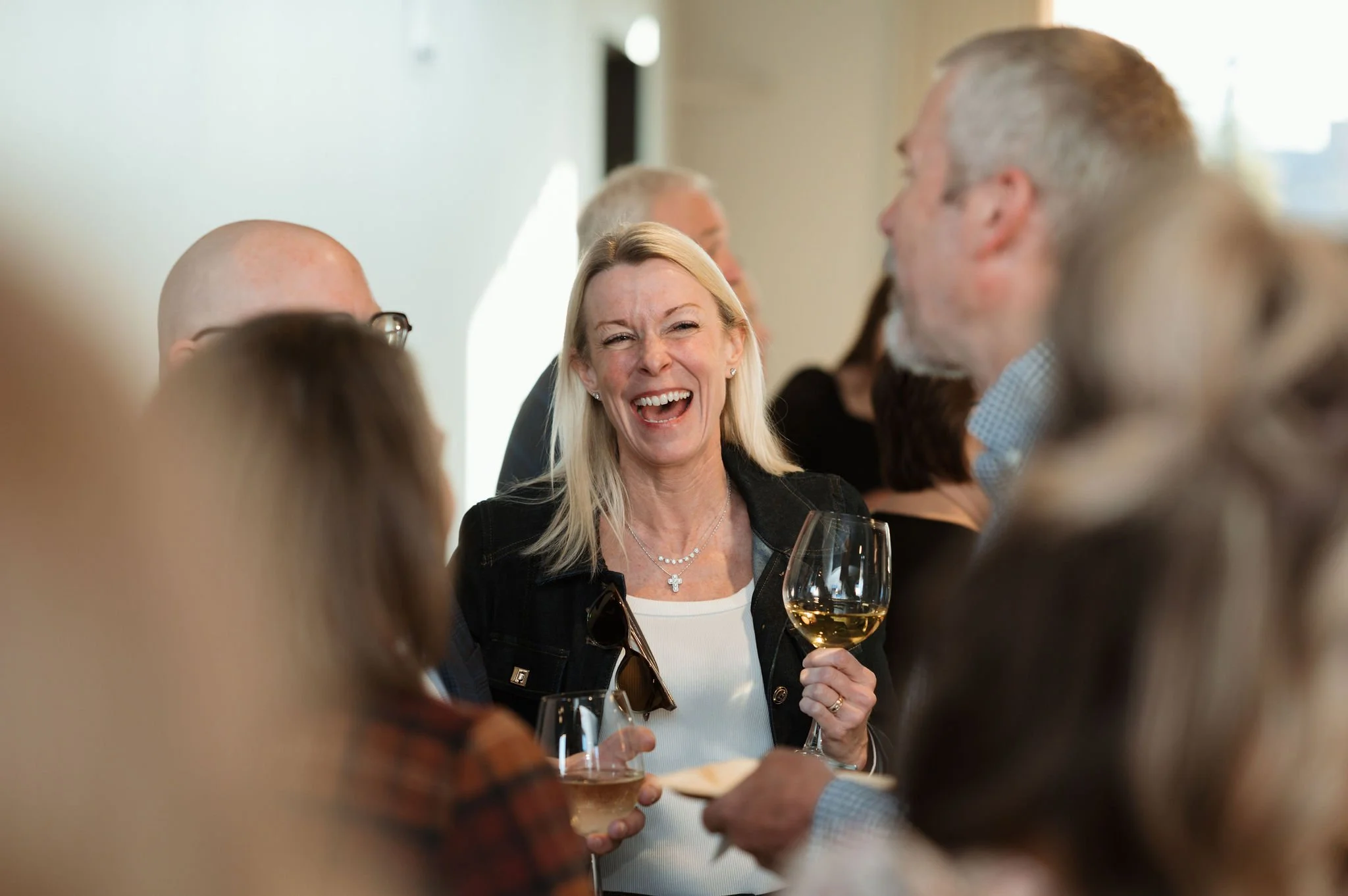 A group of people socializing at an indoor event, with a woman smiling and holding a glass of white wine.