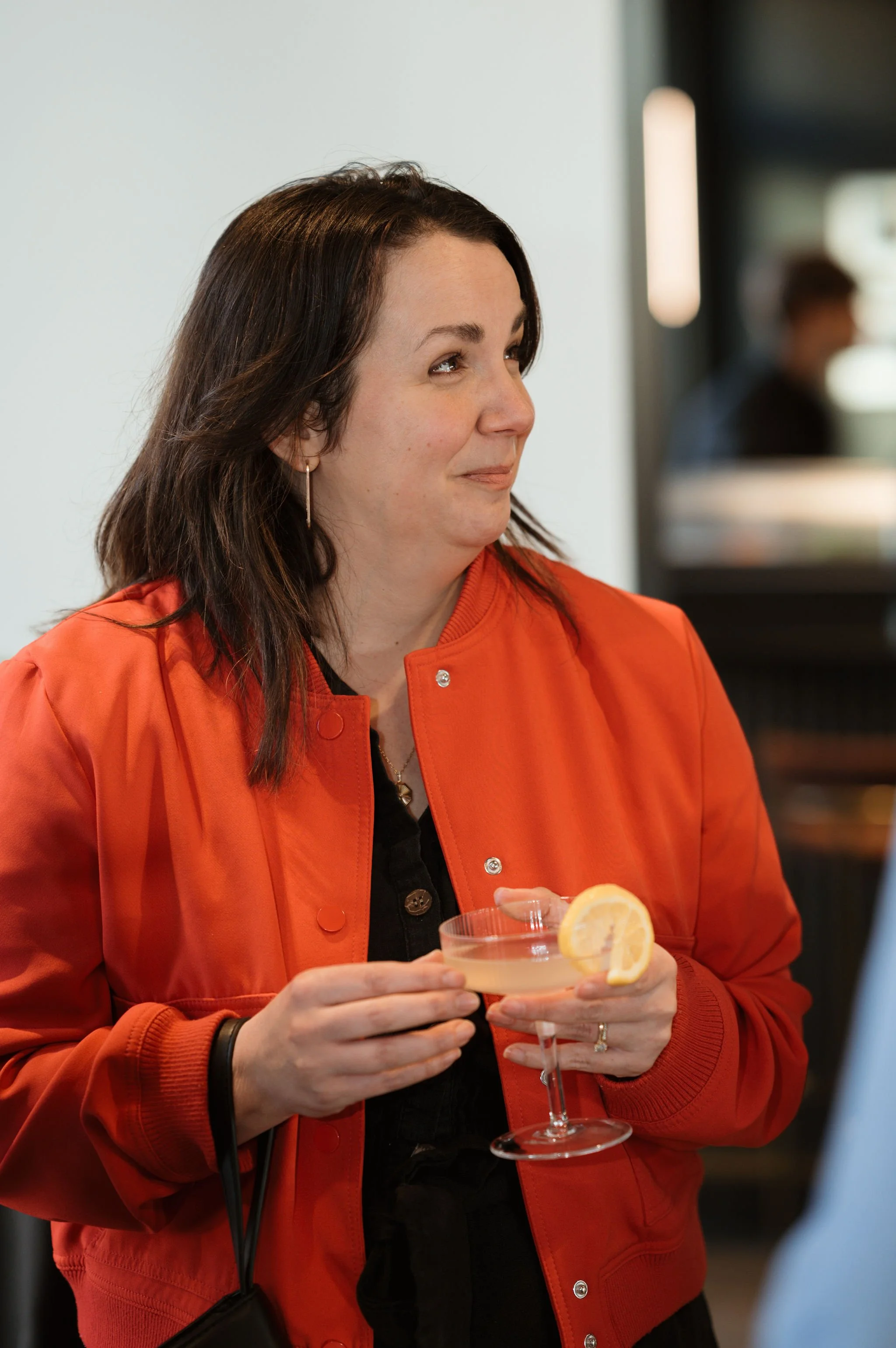 A woman with dark brown hair, wearing an orange jacket and earrings, holding a cocktail glass with a lemon garnish.