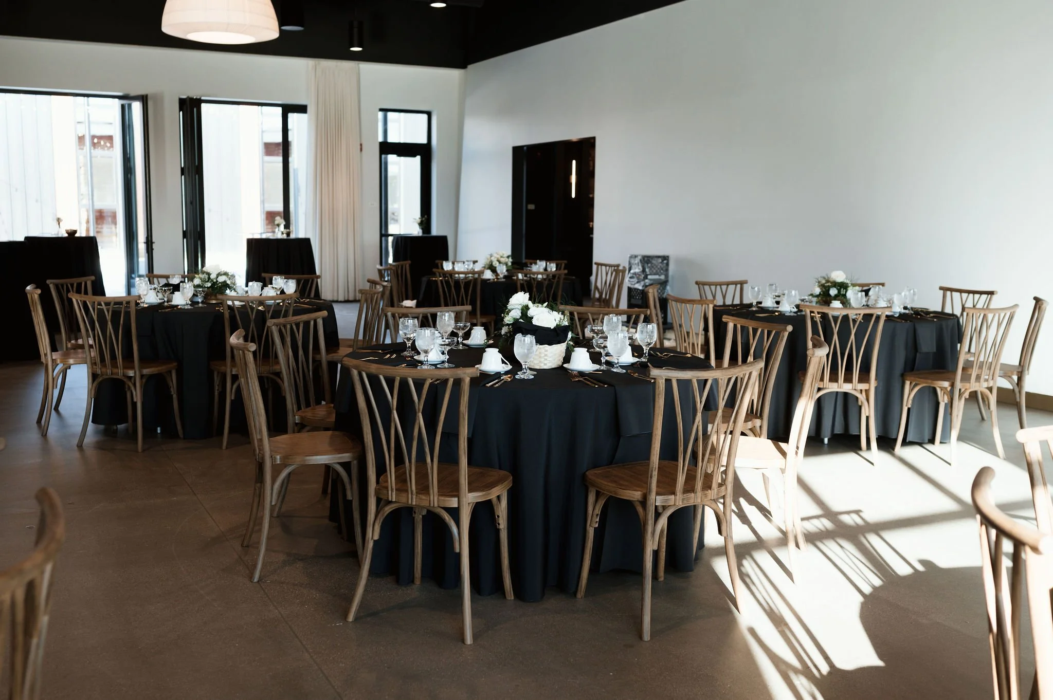 Elegant dining area with round tables covered in black tablecloths, set with glassware and white floral centerpieces. Wooden chairs surround the tables, and large windows let in natural light, casting shadows on the floor. The walls are white, with a
