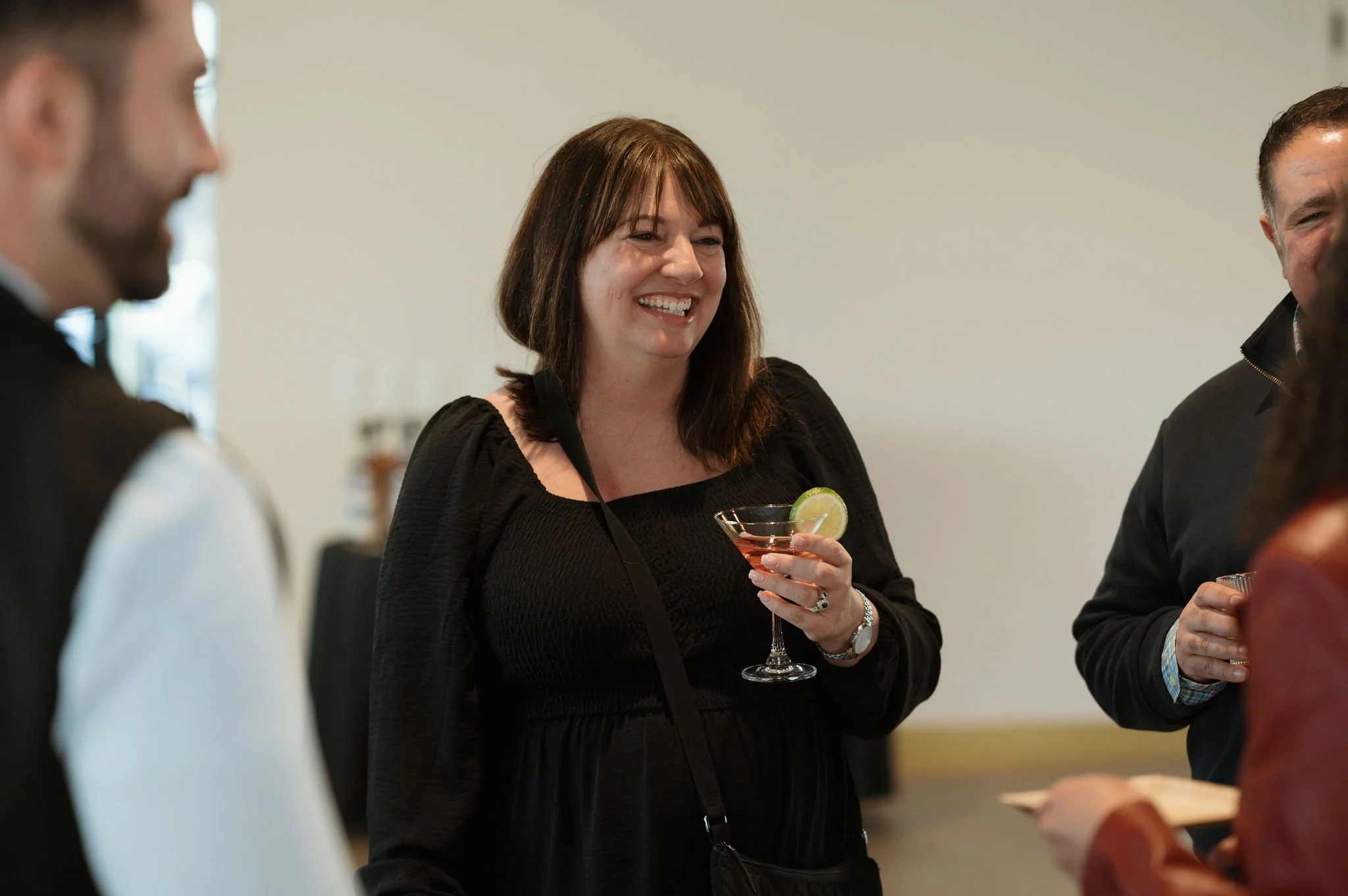 A woman with shoulder-length brown hair wearing a black dress is smiling and holding a martini glass with a lime wedge, surrounded by partially visible people at a social gathering.