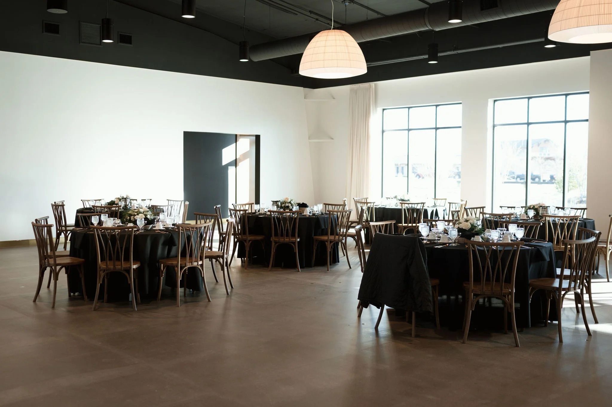 A spacious event hall with large windows letting in natural light, decorated for a formal gathering with multiple round tables covered in black tablecloths and surrounded by wooden chairs. The tables are set with glassware, and some feature floral ce