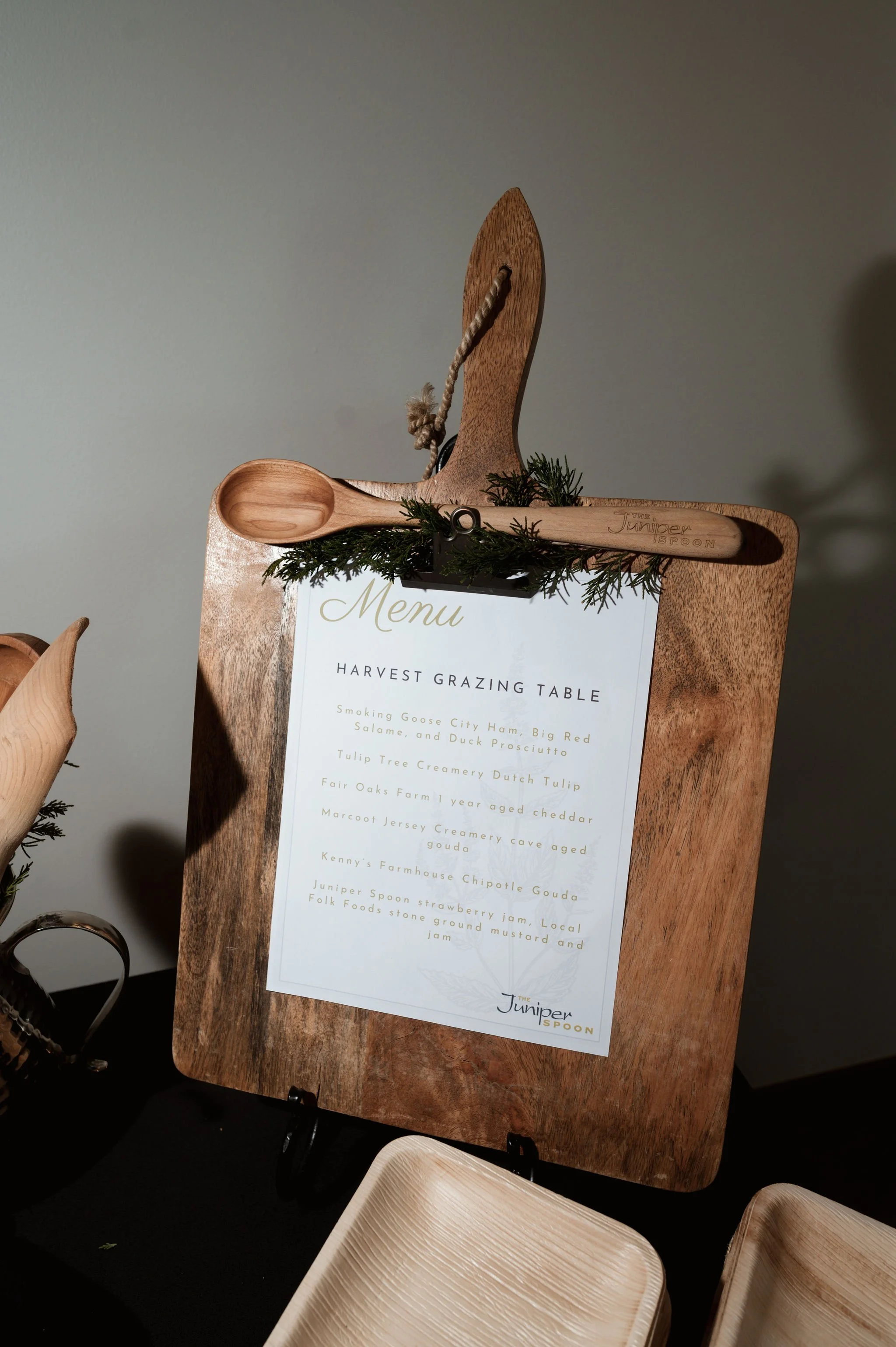 A menu on a wooden stand with a wooden spoon and greenery decoration, listing dishes at a harvest grazing table.