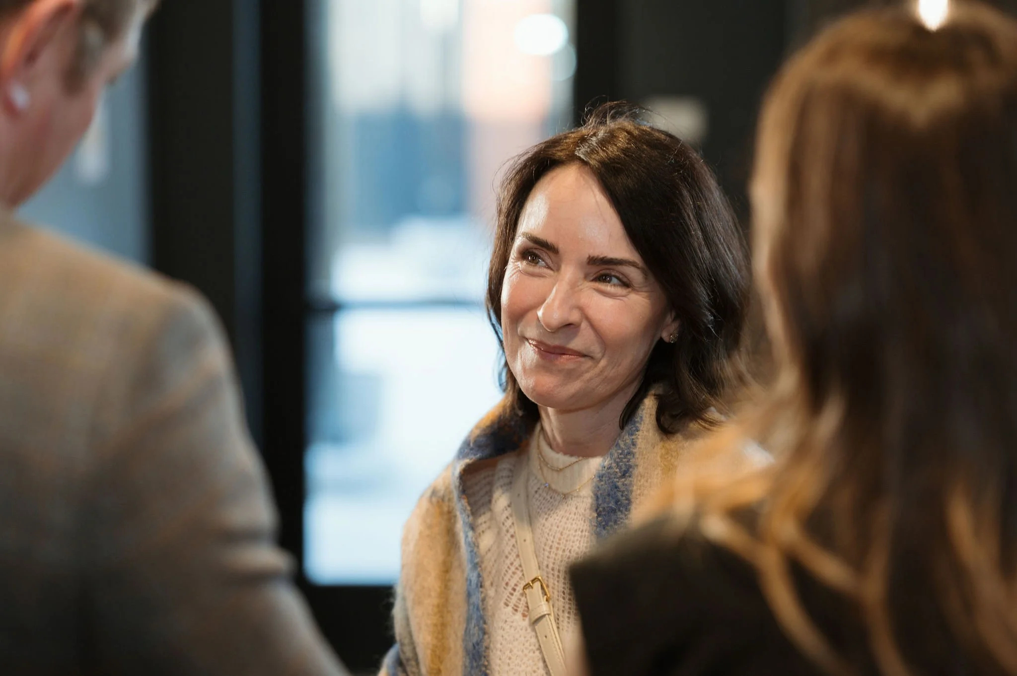 A woman with dark brown hair smiling while socializing with two people in an indoor setting.