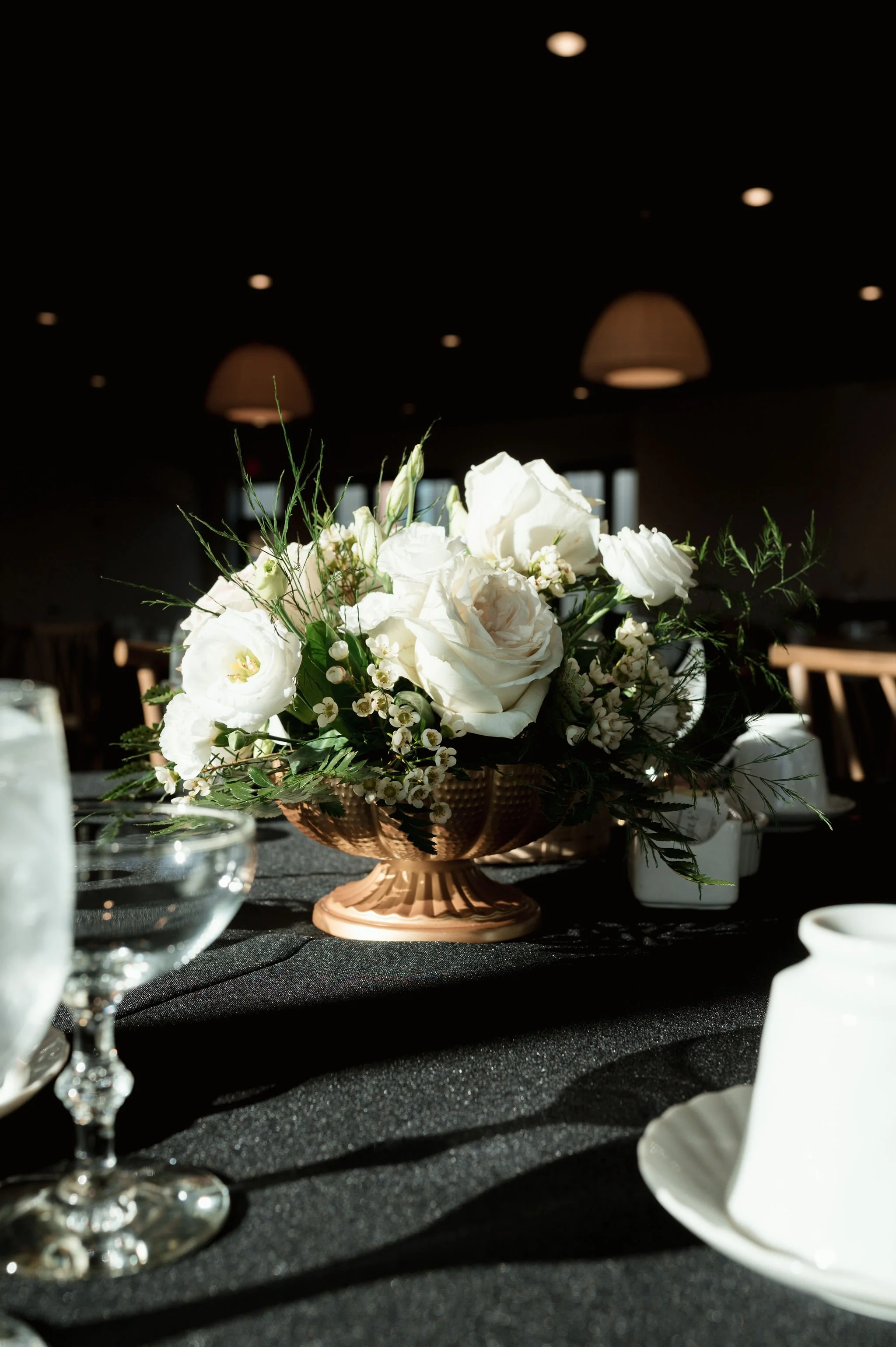 A floral centerpiece with white roses, lisianthus, and greenery in a copper-colored pedestal vase on a dark tablecloth, surrounded by glassware and tableware under dim lighting.