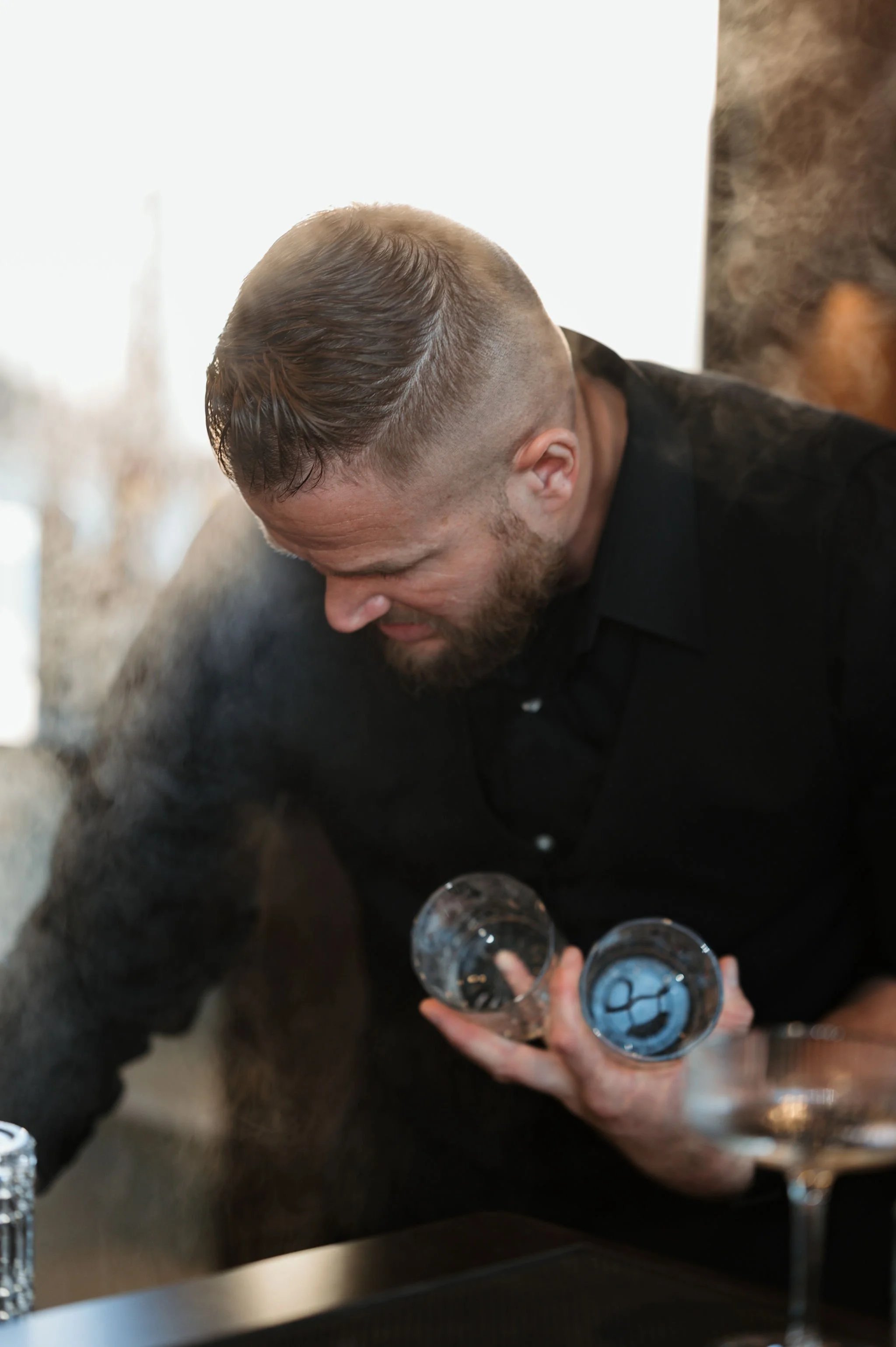 A man with a neatly styled haircut, wearing a black shirt, is holding two glasses of water. He is leaning over a surface, possibly at a social gathering or event, with other people partially visible around him.