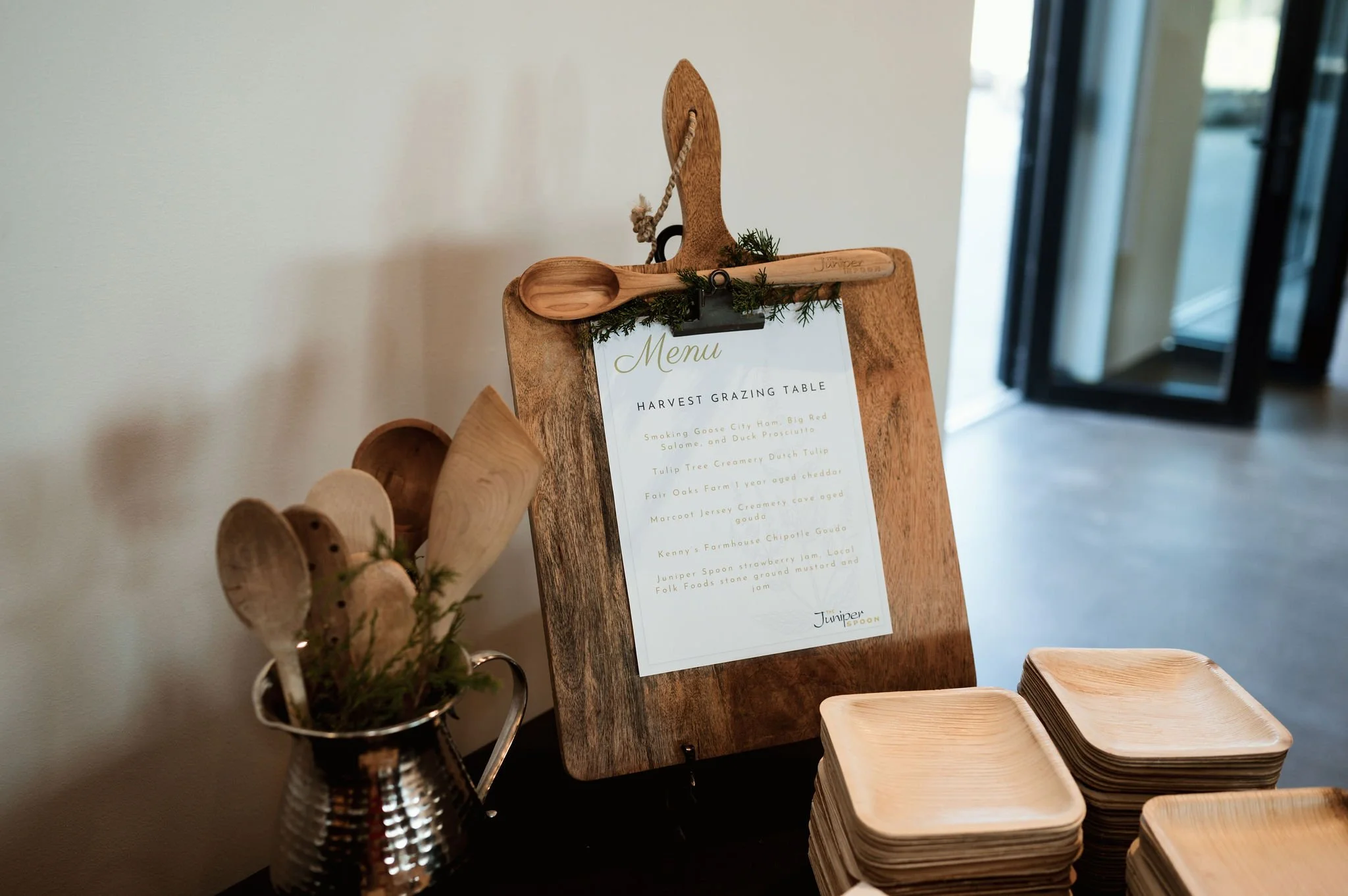 A wooden menu board with a small wooden spoon attached, decorated with greenery, next to a metal container holding wooden utensils and a stack of square wooden plates.
