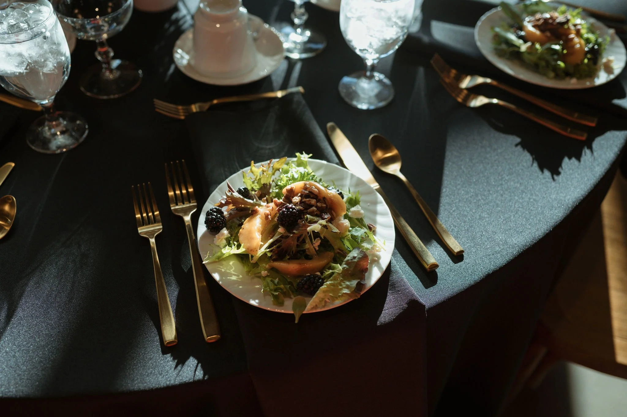 A table set for a meal with gold cutlery, glasses of water, and a salad on a white plate. The table has a dark tablecloth and appears to be in a dimly lit setting.