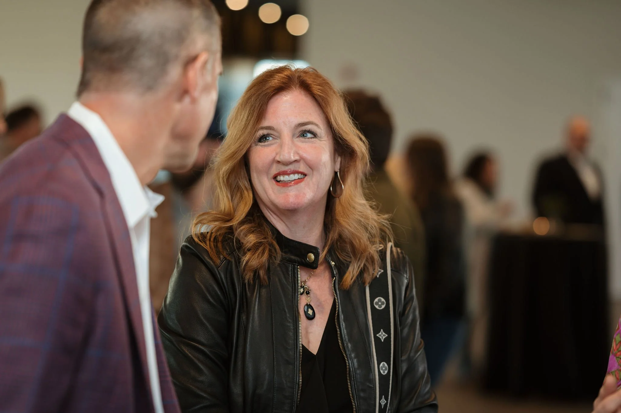 A woman with long red hair wearing a black leather jacket is talking to a man with short hair wearing a checkered suit jacket. They are in a crowded room with blurred people in the background.